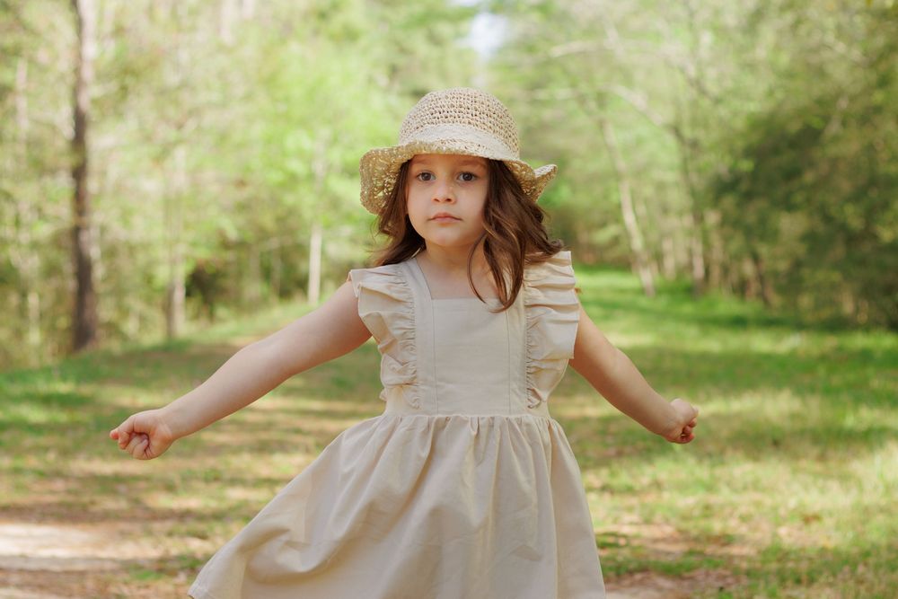 A young woman in a beige dress and sun hat stands in a lush, green forest with sunlight filtering through the trees.