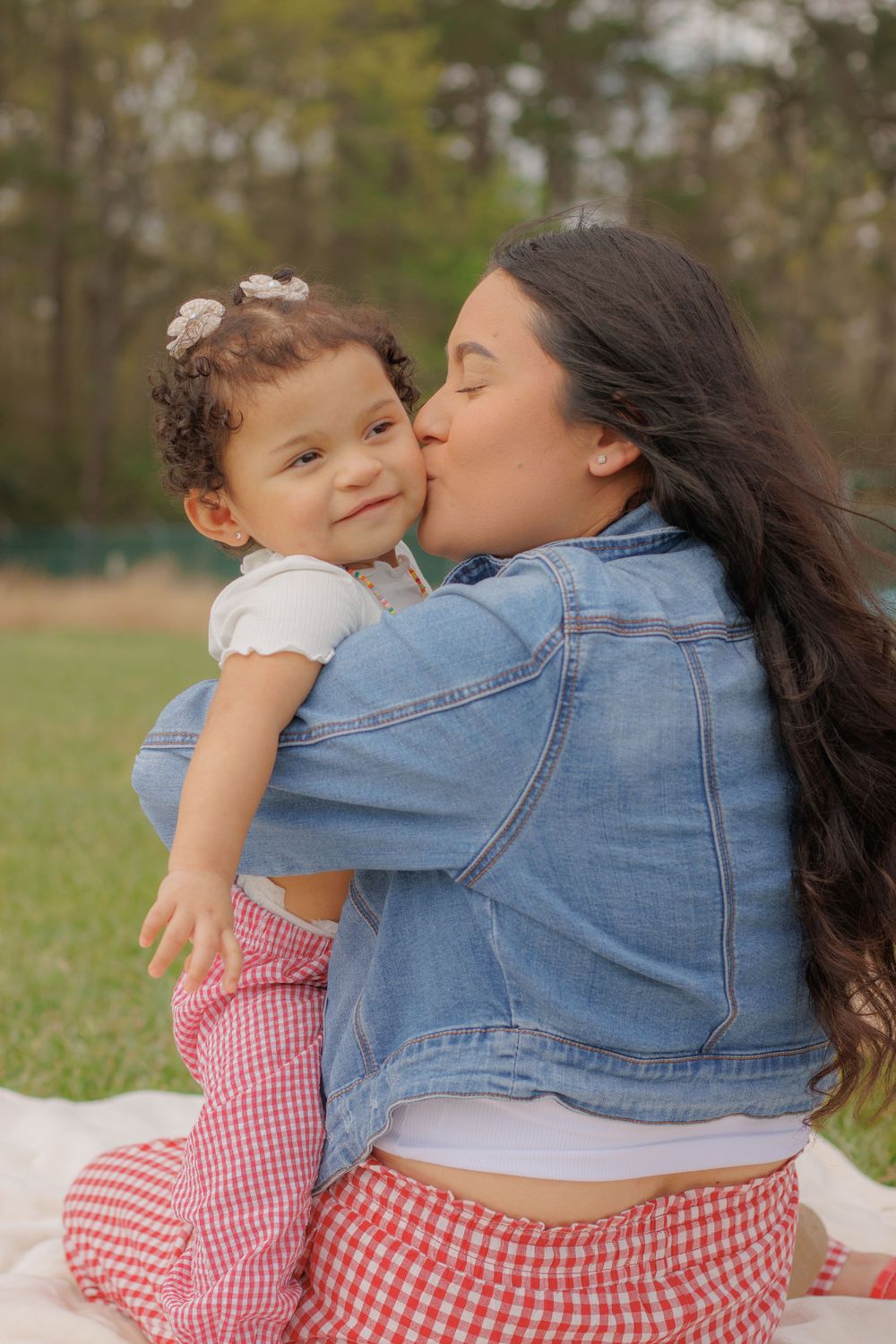 A mother embraces and kisses her young daughter in a grassy outdoor setting.