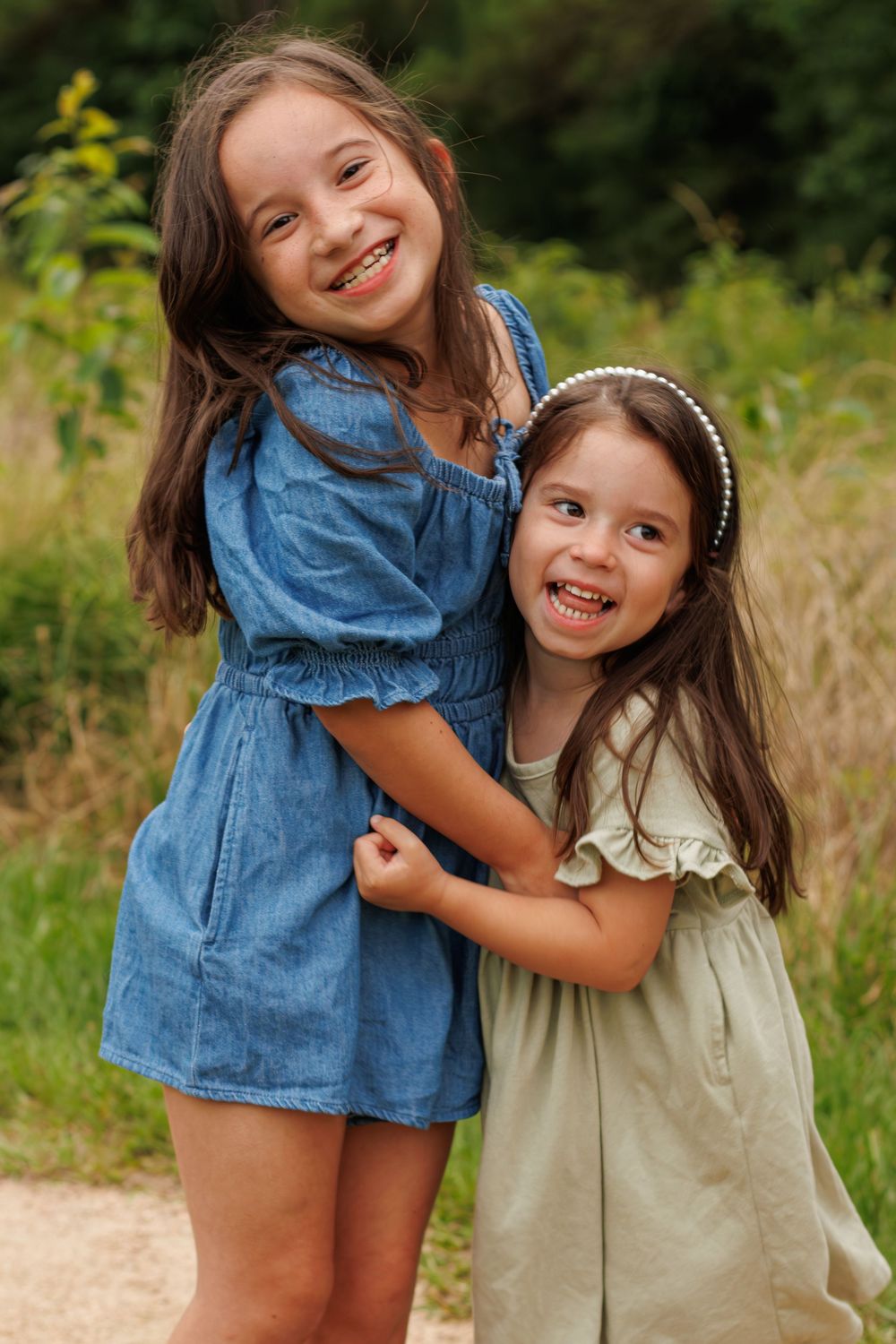 Two young girls in summer dresses smile and hug outdoors, one in denim and one in sage green with a pearl headband.