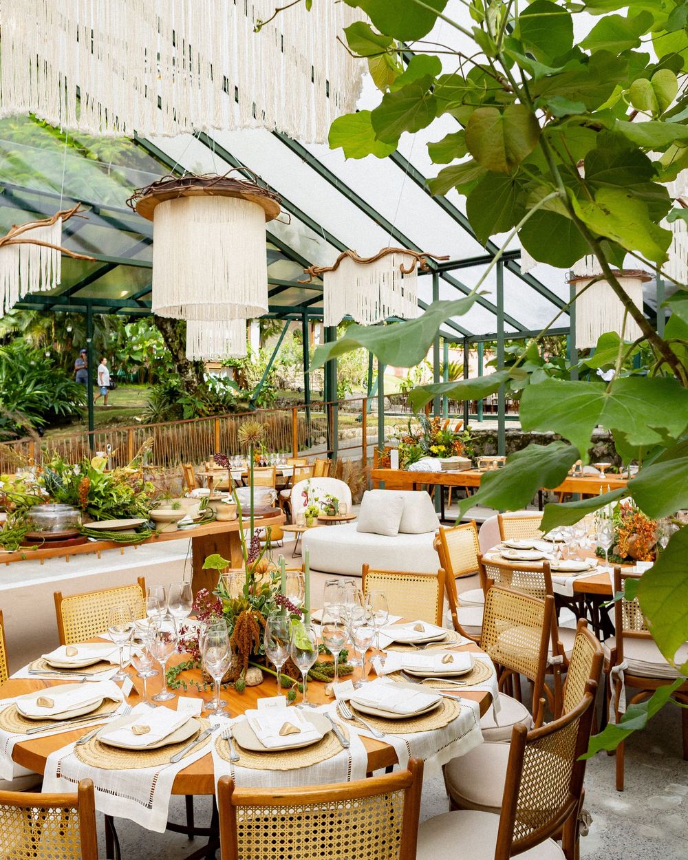 Elegant greenhouse restaurant interior with rattan chairs, white cushions, and lush greenery overhead creating a botanical dining space.