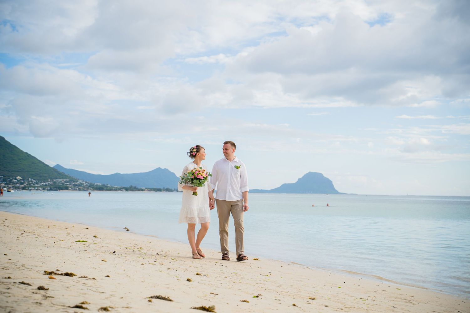 A couple in white attire walks along a scenic tropical beach with mountains and ocean in the background. Shot by Luminair Studio