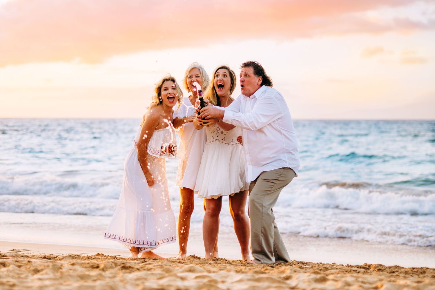 A family wearing white outfits poses together on a beach at sunset with ocean waves in the background.