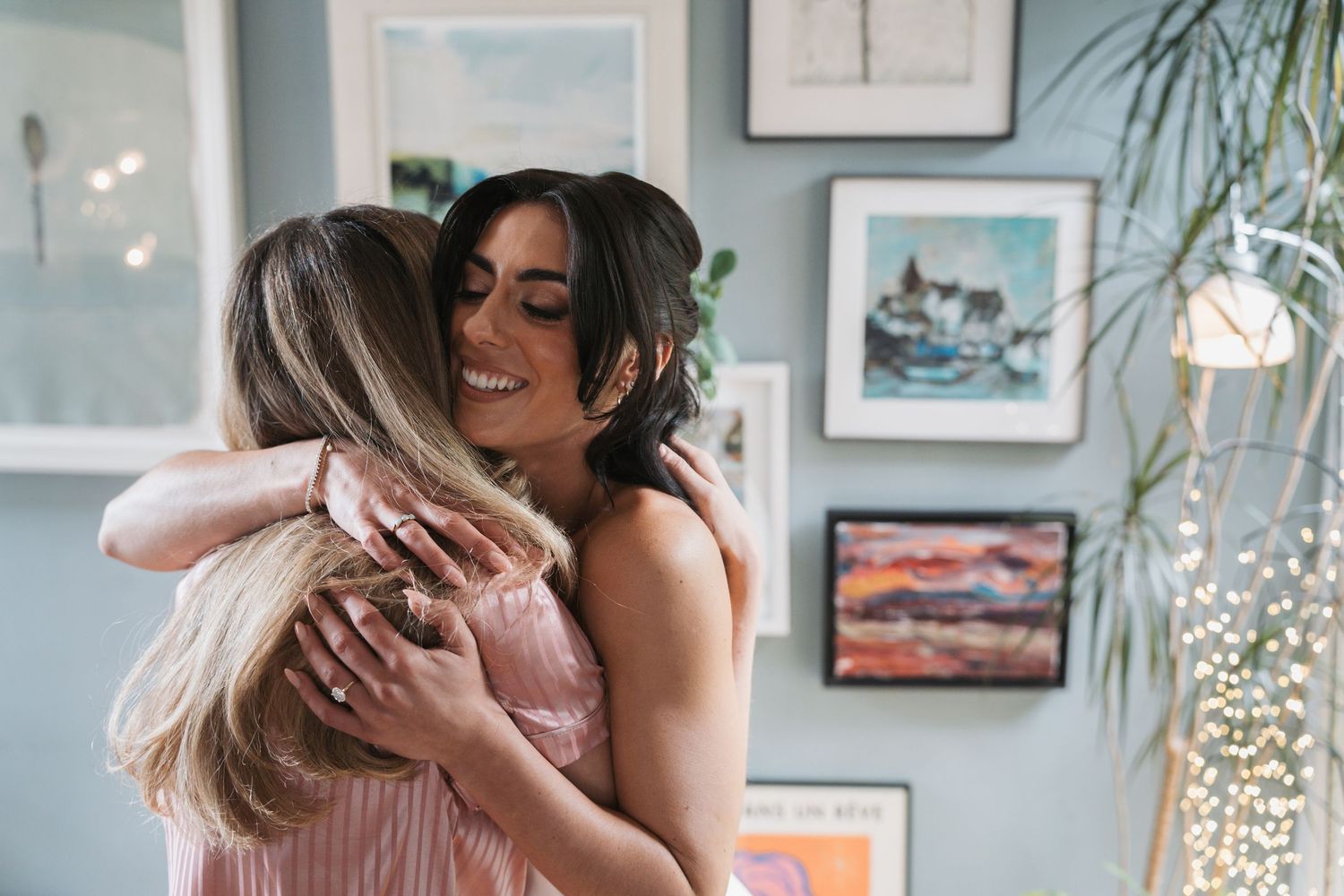 Two people share a tender embrace in a bright room decorated with framed artwork and plants.