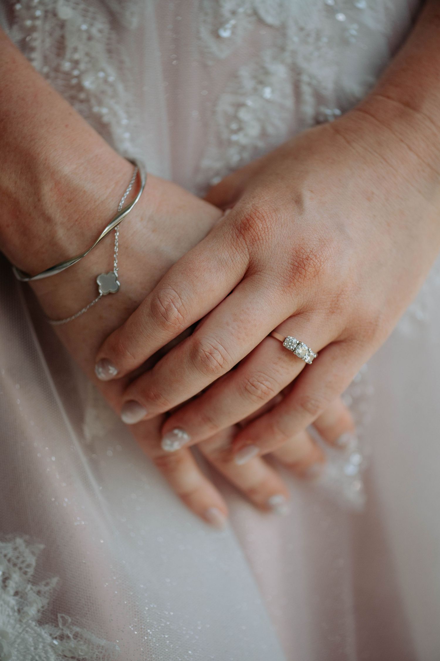 Close-up view of delicate hands wearing engagement rings against a white lace dress background.