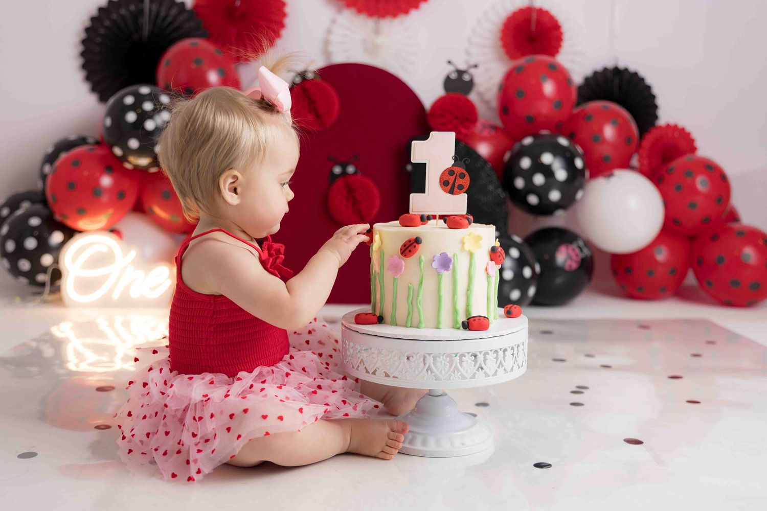 A cake smash photo session setup with red and black balloons, white cake, and pink tulle decorations.