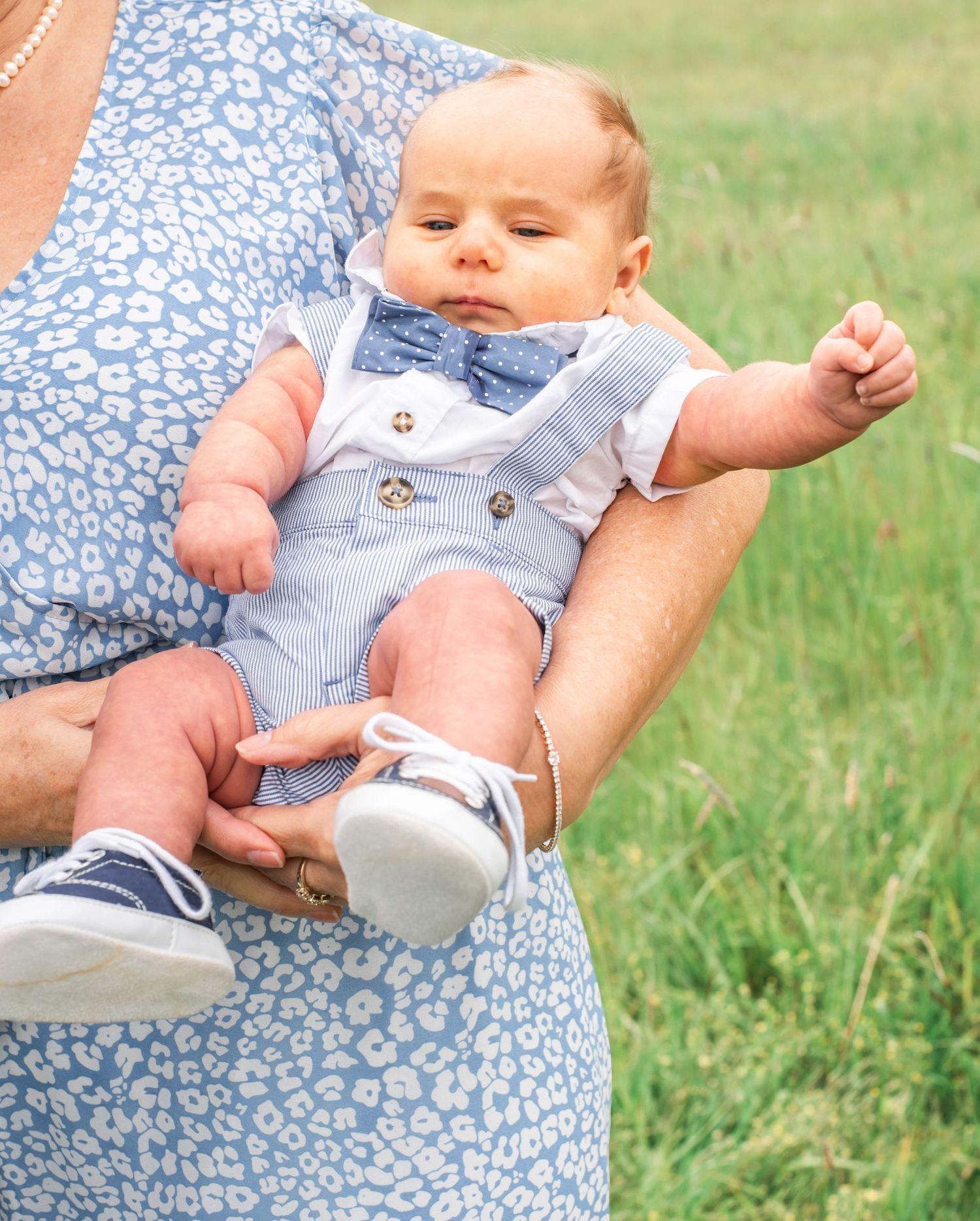 Baby in light blue overalls being held against floral print fabric in outdoor grassy setting.