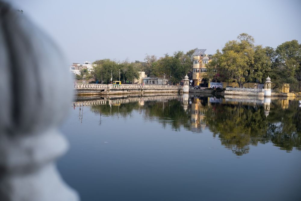 Serene lake view with ancient temple structures and lush trees reflecting on calm water in Udaipur, India.