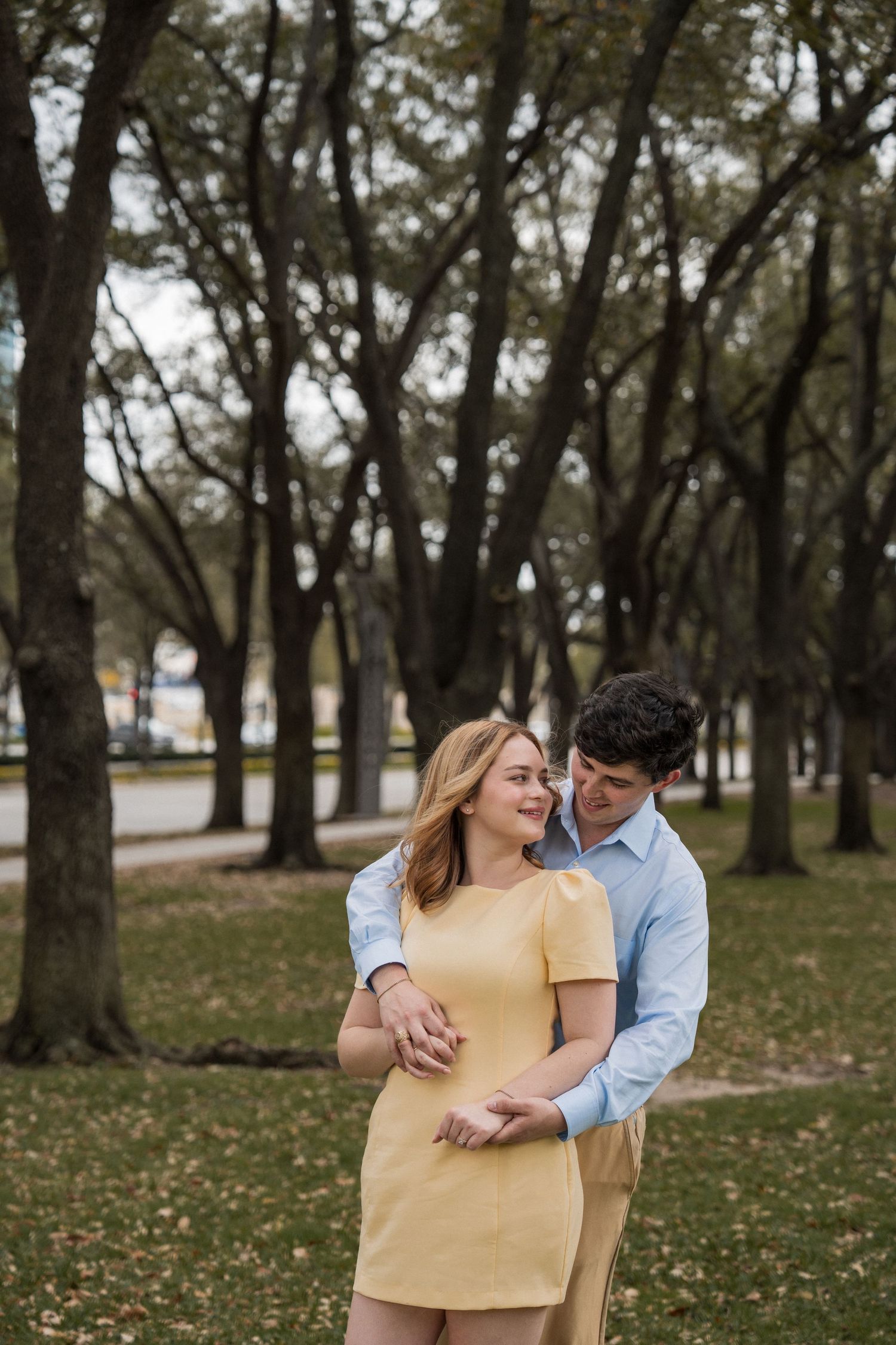 A romantic couple shares tender moments in a park setting with trees and fallen leaves in the background.