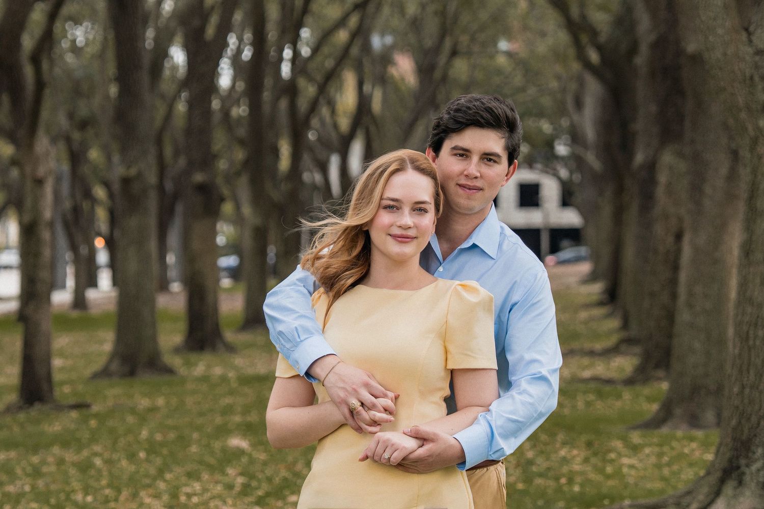 A couple in casual attire embraces while walking through a tree-lined path on a spring day.