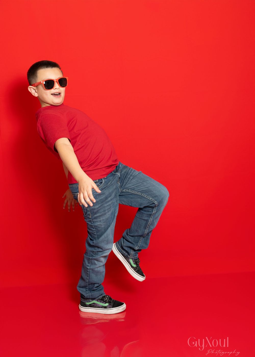 Young child in red shirt and jeans strikes a playful dance pose while wearing sunglasses against a bright red background.