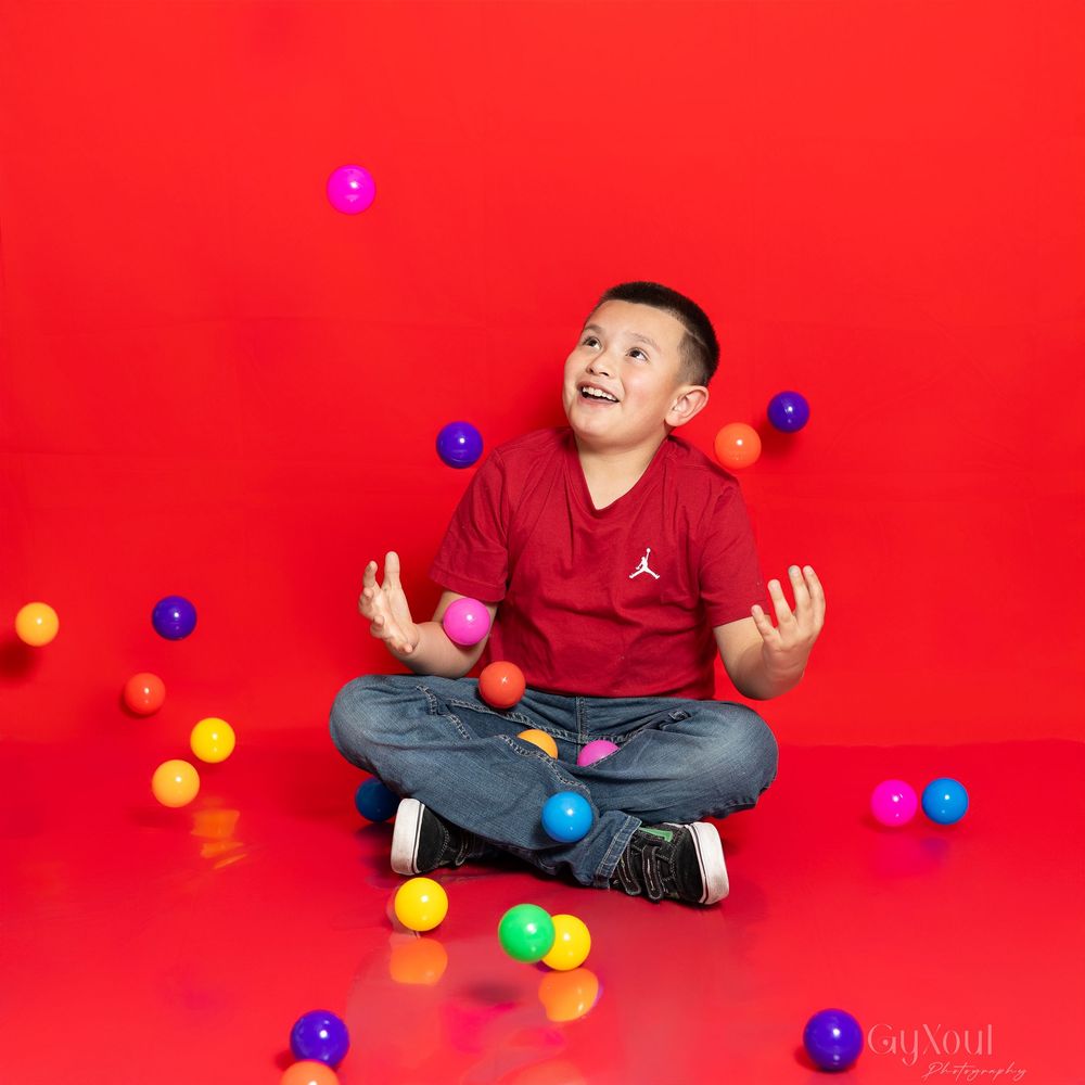 Young person in red shirt sitting cross-legged juggling colorful balls against vibrant red background.