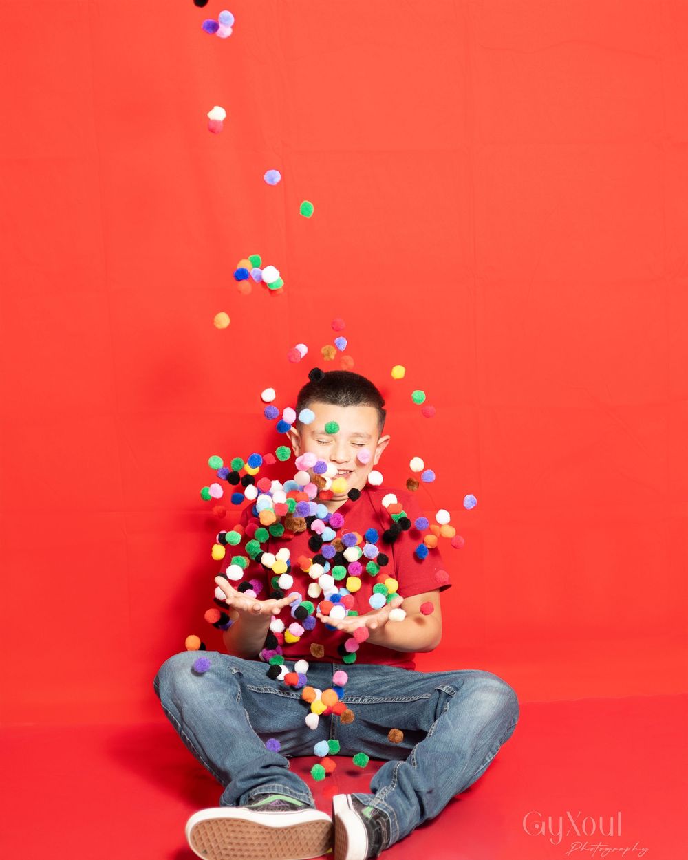 Colorful confetti falls around a person in jeans sitting cross-legged against a bright red background.