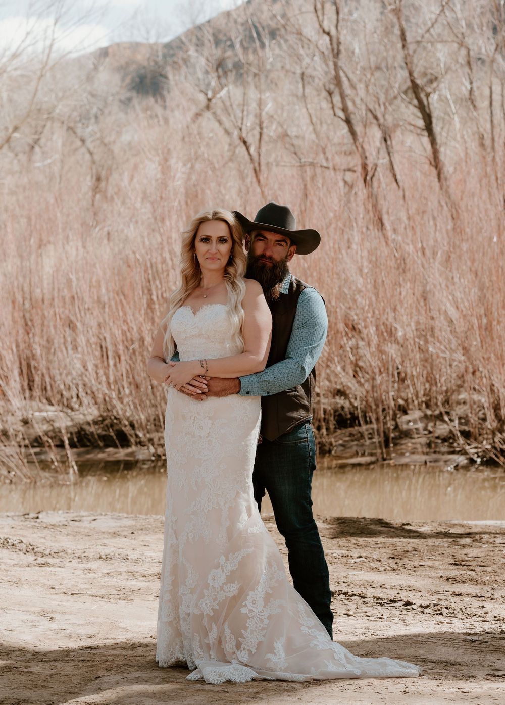A couple embraces in a romantic moment by a sunlit riverbank in sepia tones.