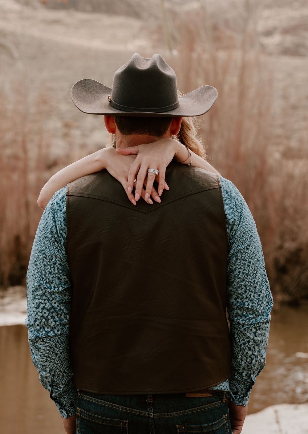 A couple in casual attire embraces from behind while sitting outdoors against a rustic backdrop.