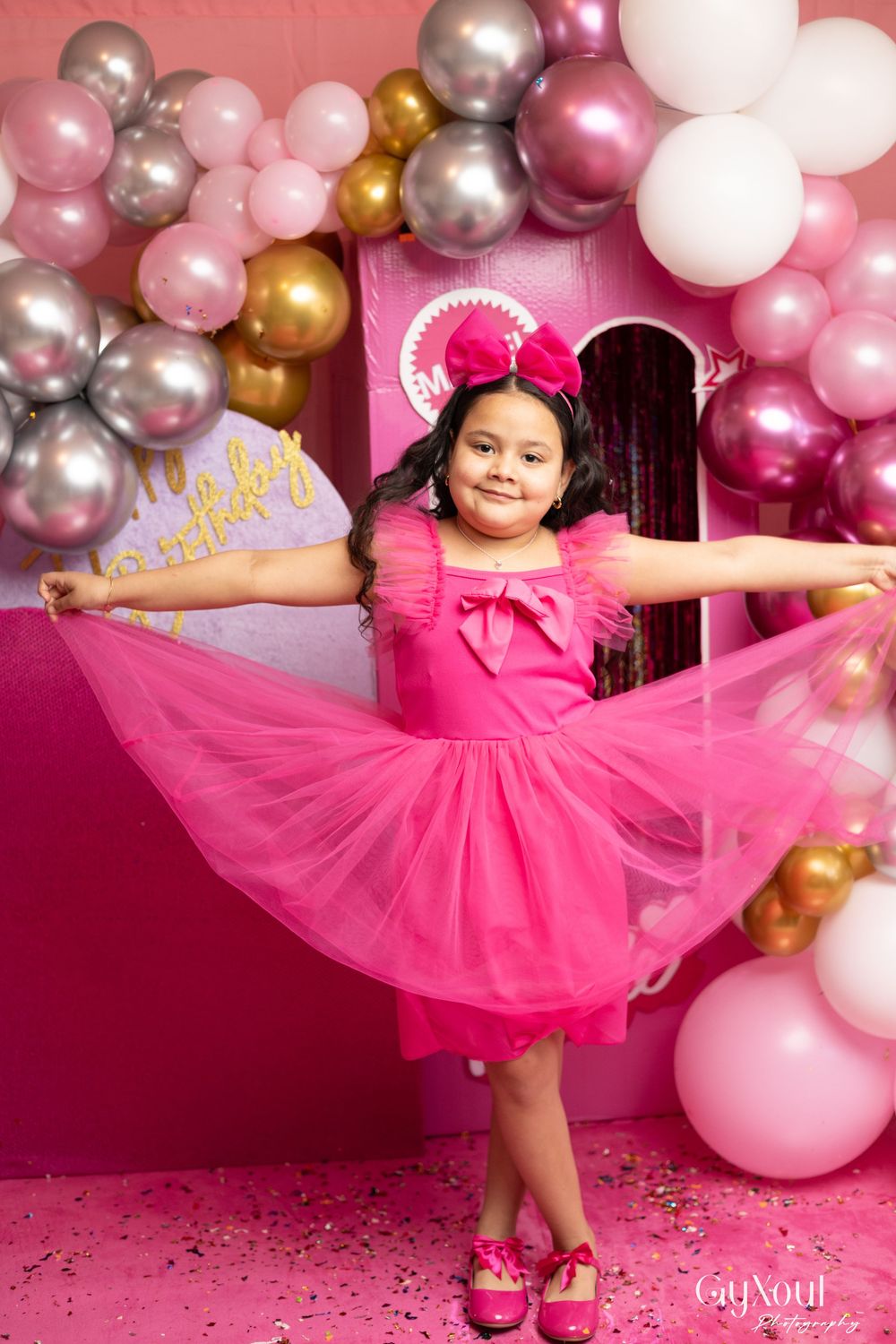 A child twirls in a pink tulle dress against a backdrop of metallic balloons at a Barbie-themed photoshoot.