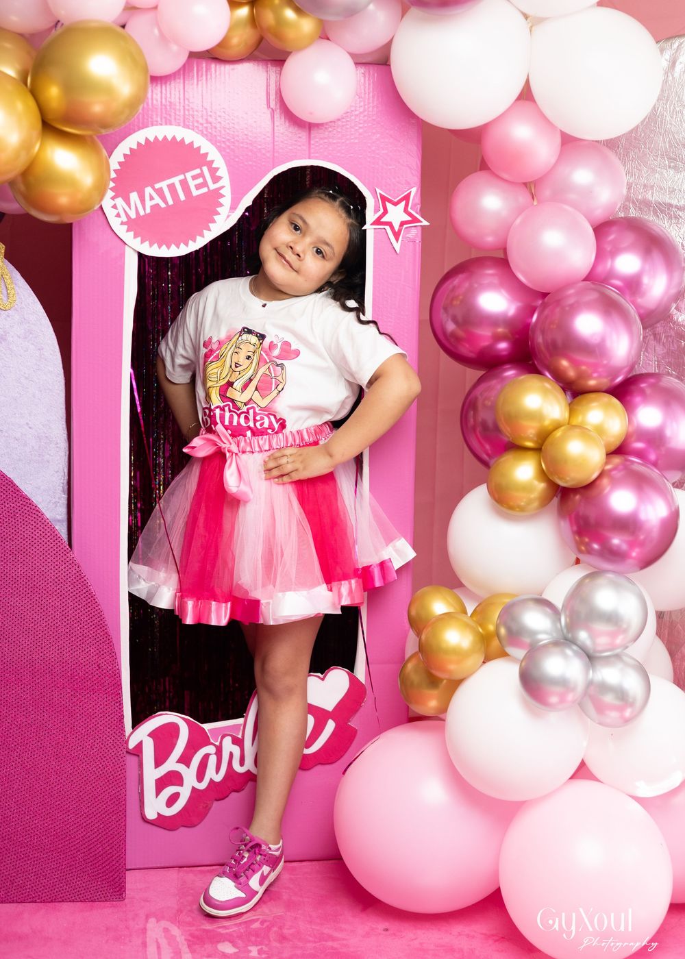 A child poses inside a life-sized pink Barbie doll box decorated with gold and pink balloon garlands.
