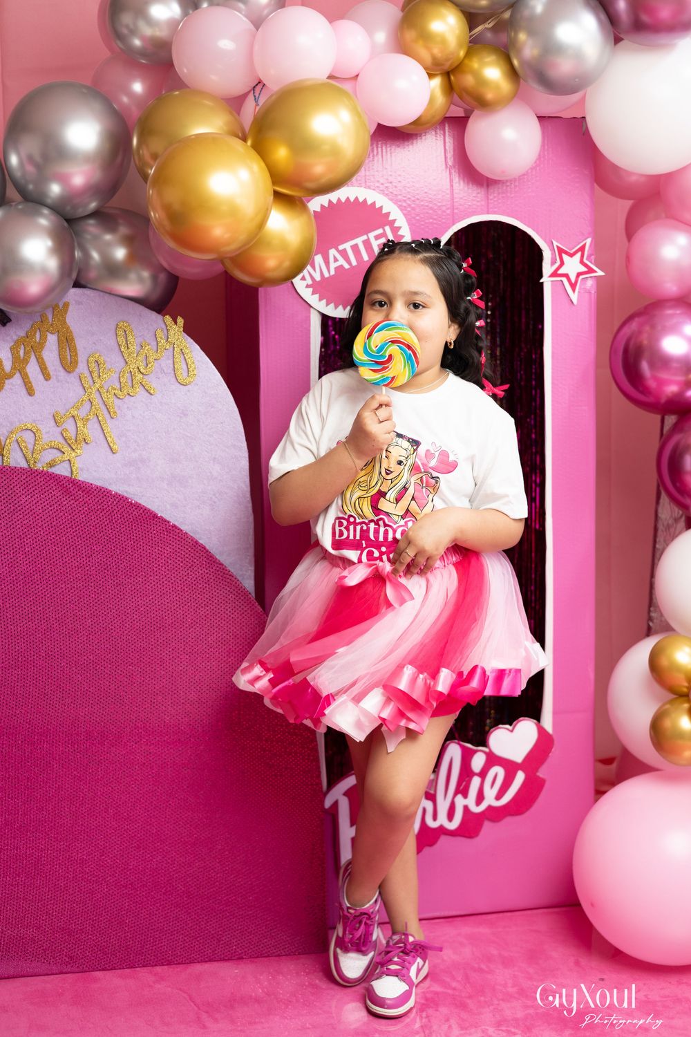 A birthday party setup with pink and gold balloon arch, featuring a pink backdrop and someone holding a lollipop.