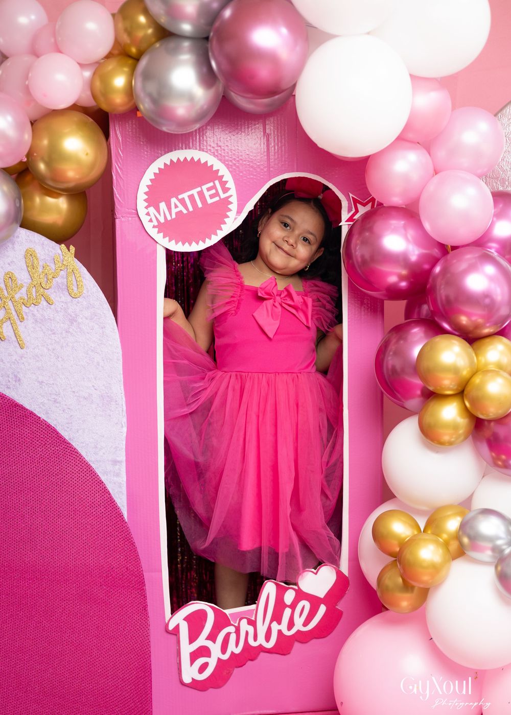 A child stands in a large pink Barbie-themed box wearing a bright pink dress with balloon decorations.
