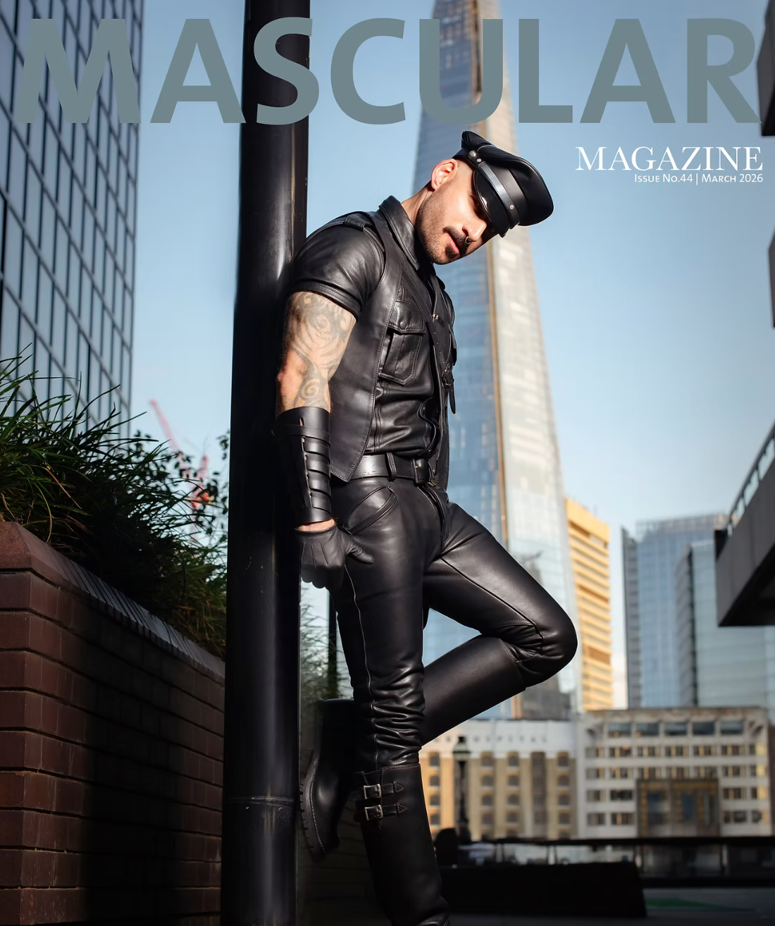 Man in full black leather outfit and cap posing against a pole with city skyline, Mascular Magazine cover.