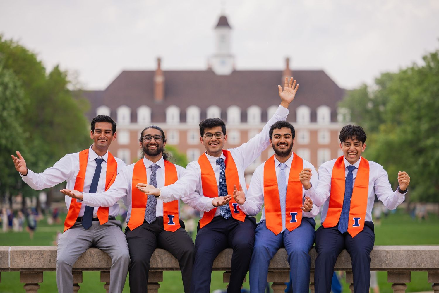 Group of five UIUC graduating students with arms up celebrating in front of the Illini Union