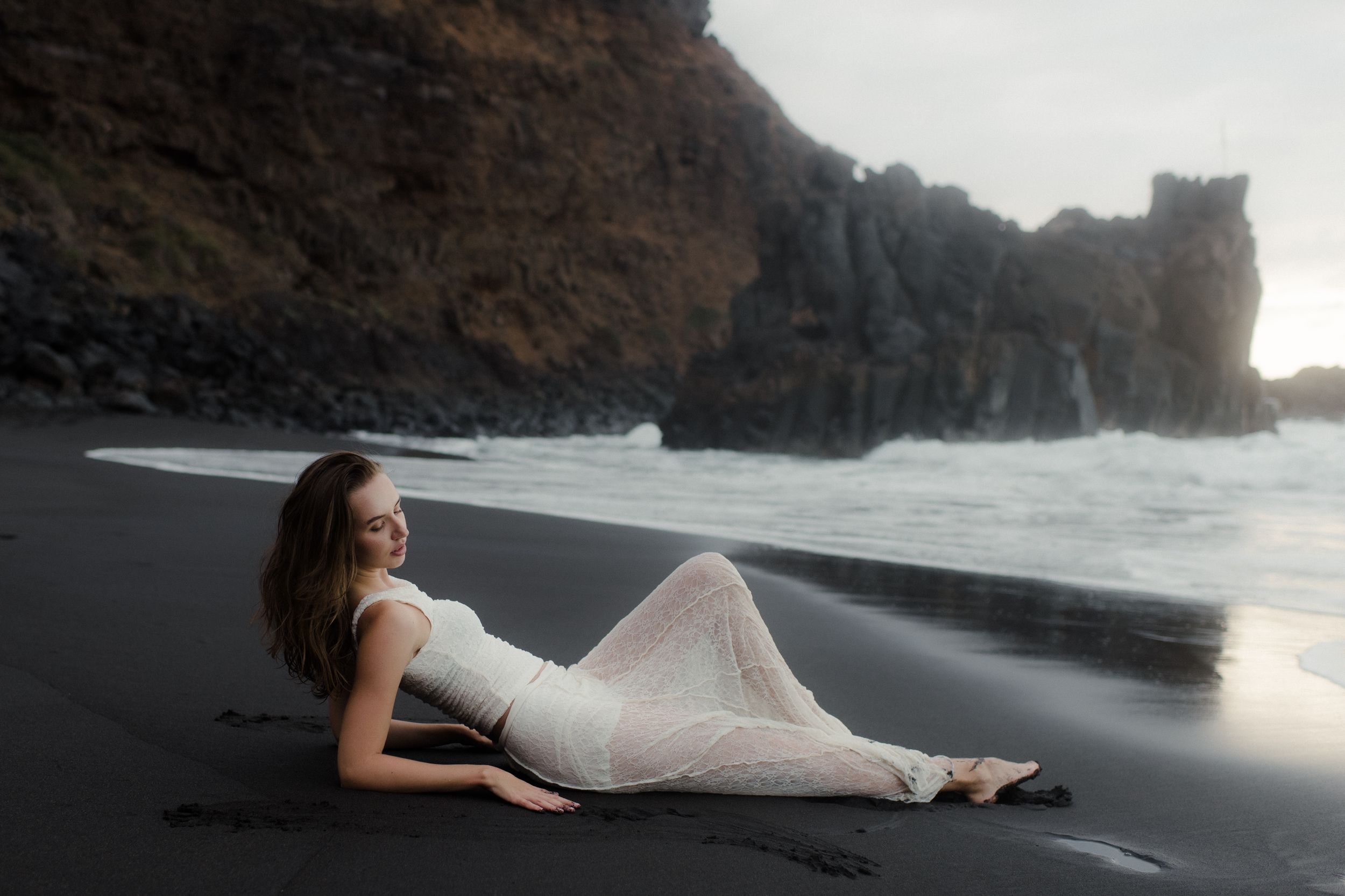 A woman in a white dress reclines on a black sand beach with dramatic cliffs and ocean waves in the background.
