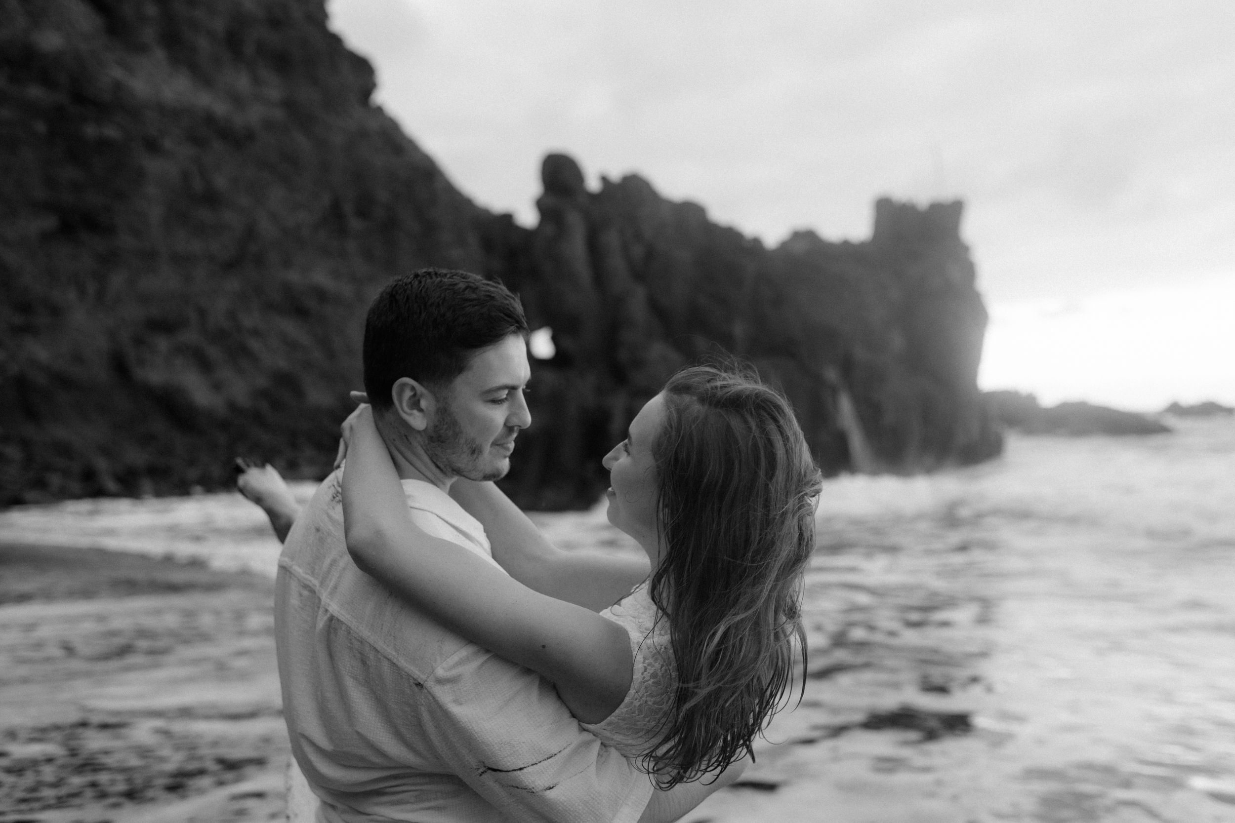 A couple embraces on a scenic beach with waves crashing against rocky cliffs in black and white.