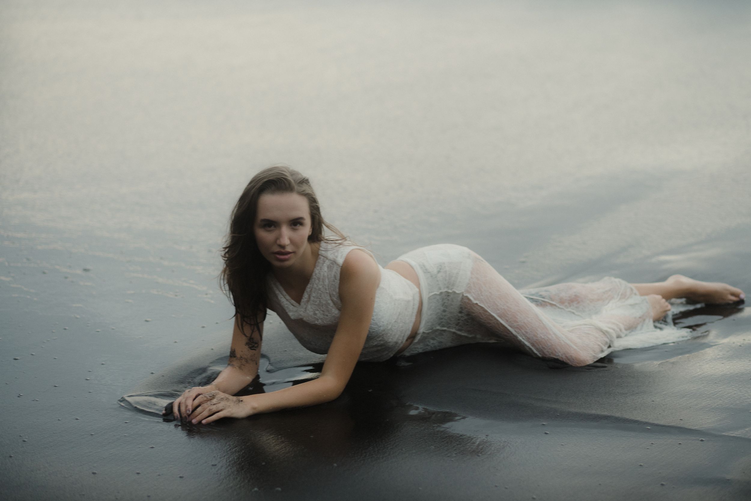 A person in a white dress lies gracefully on a misty beach during an ethereal photoshoot.