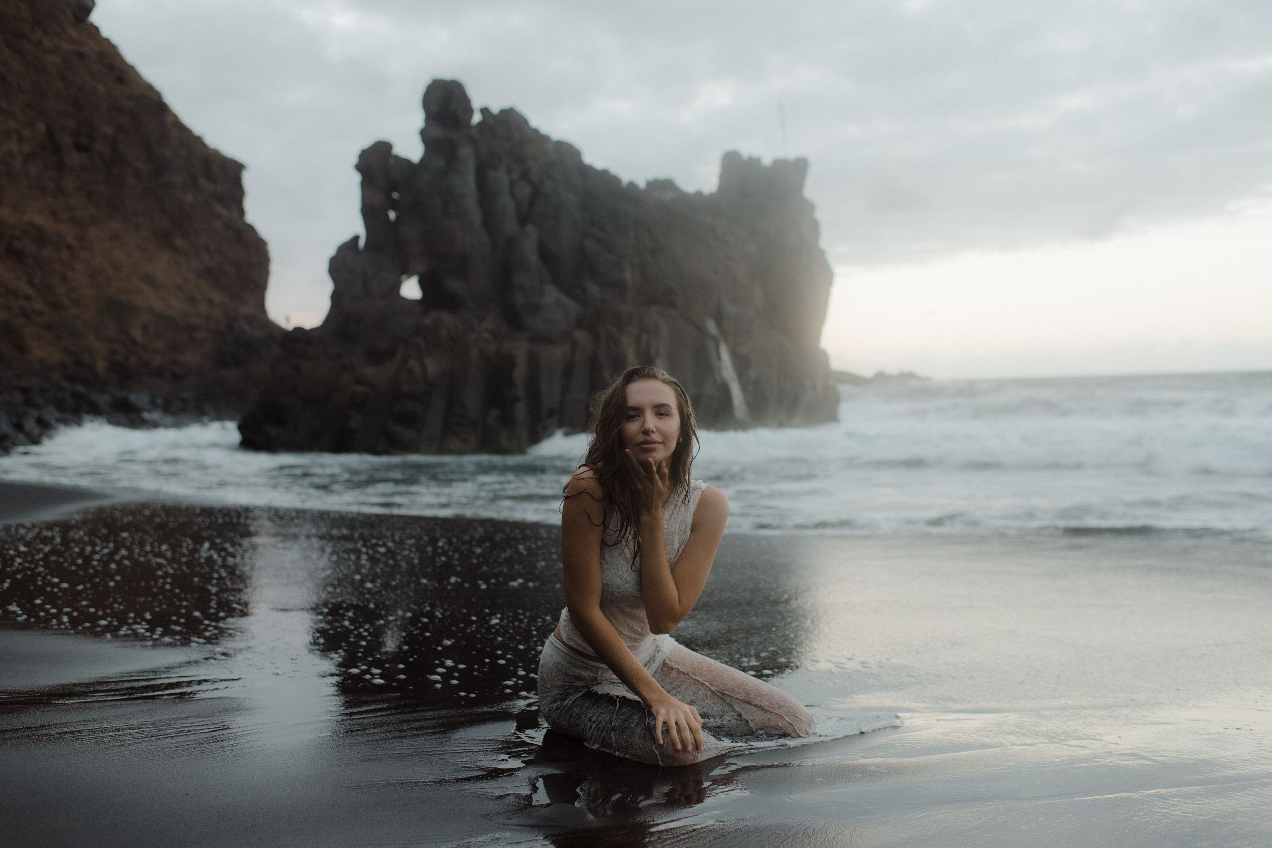 Couple exploring a misty black sand beach with dramatic coastal cliffs and crashing waves in moody weather.