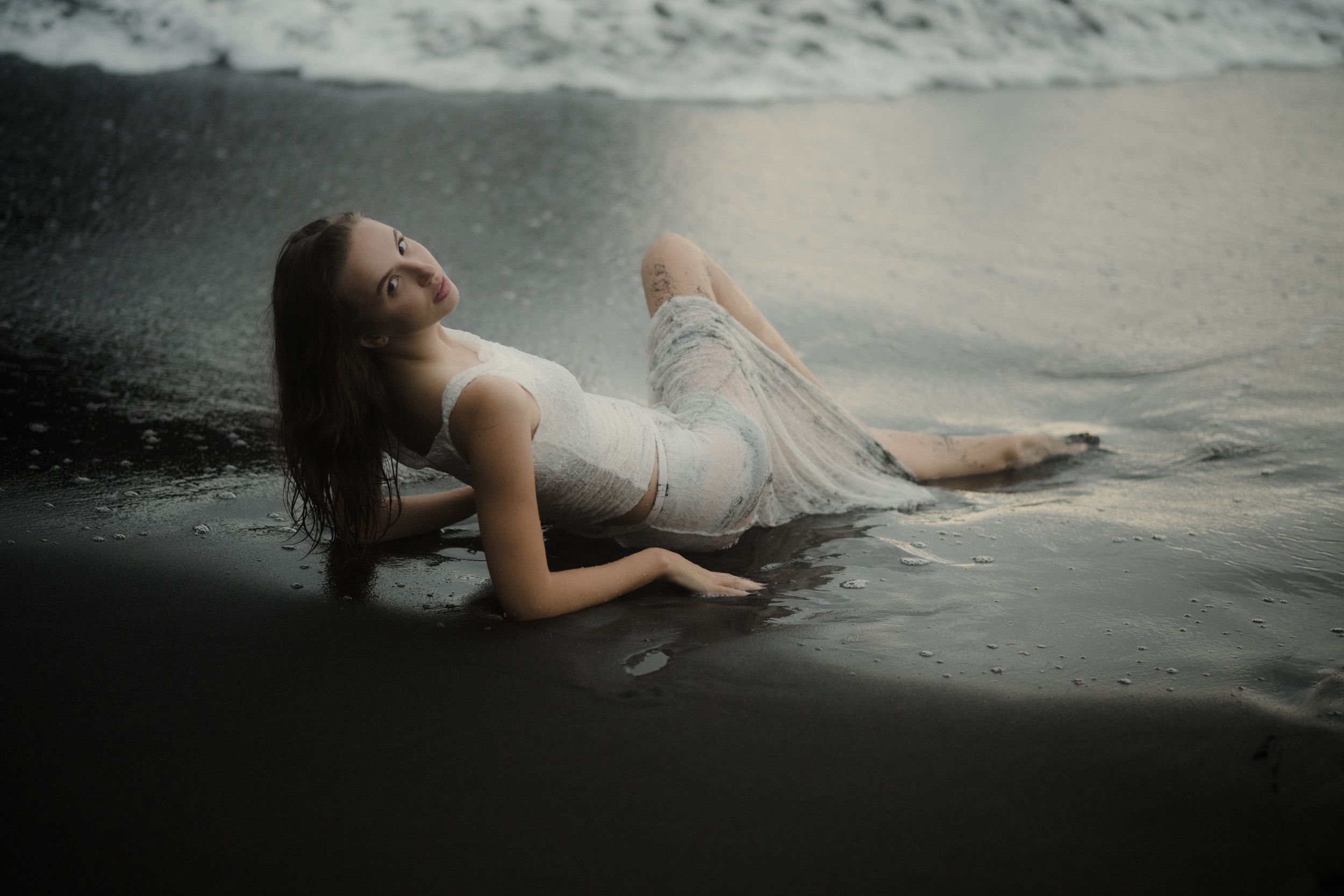 A dreamy beach photoshoot features a person in white reclining on black sand near crashing waves.