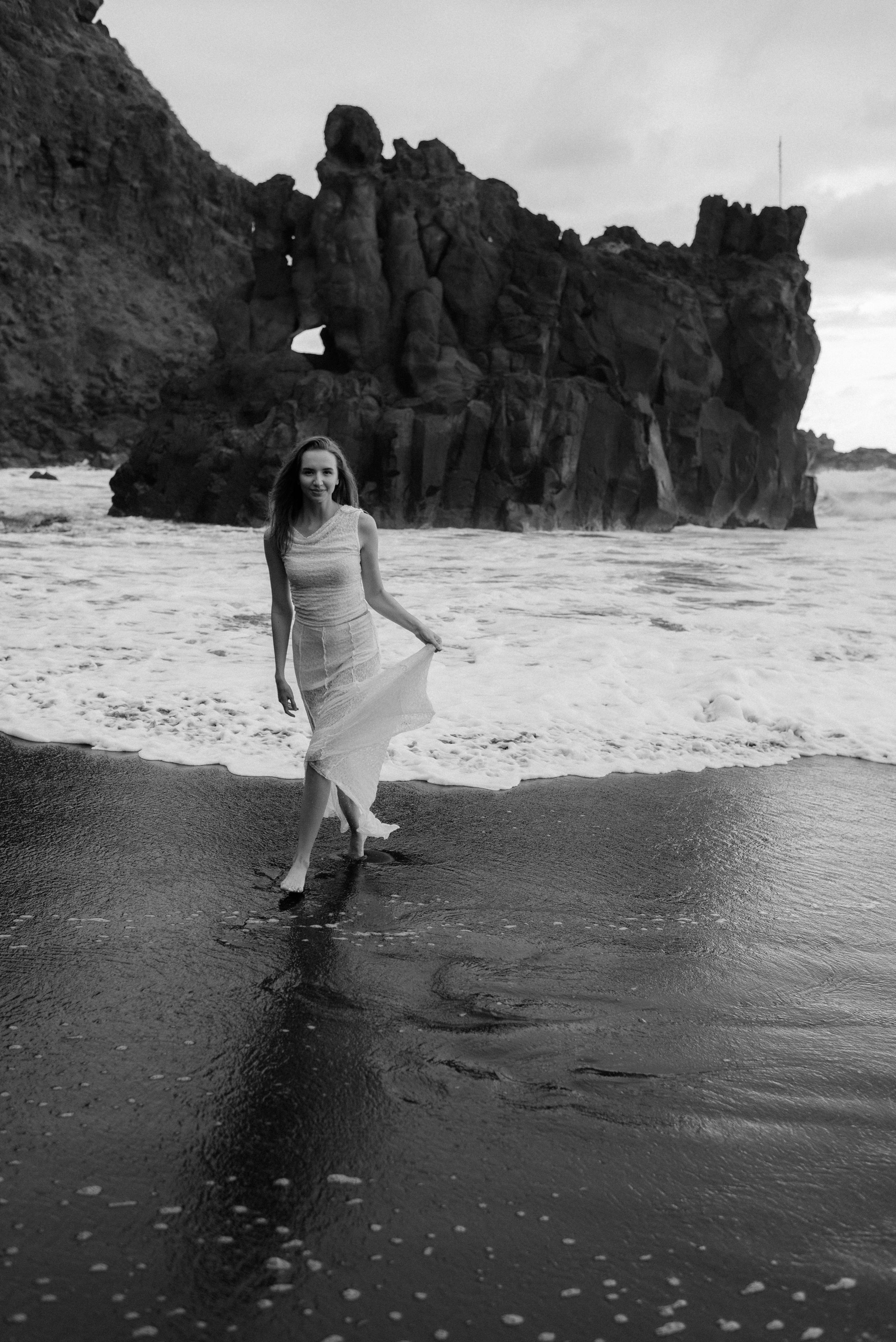 A person in white dress walks along a dramatic rocky beach shoreline in black and white photography.