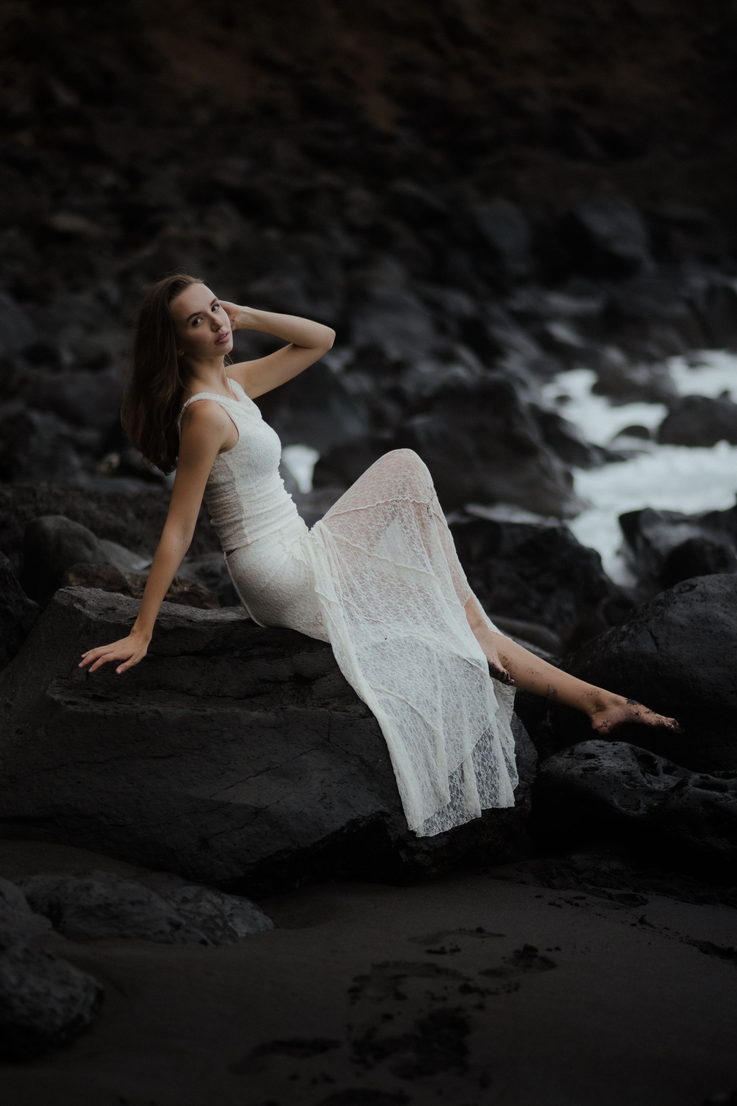 A person in a white lace dress poses dramatically on dark volcanic rocks near the ocean.