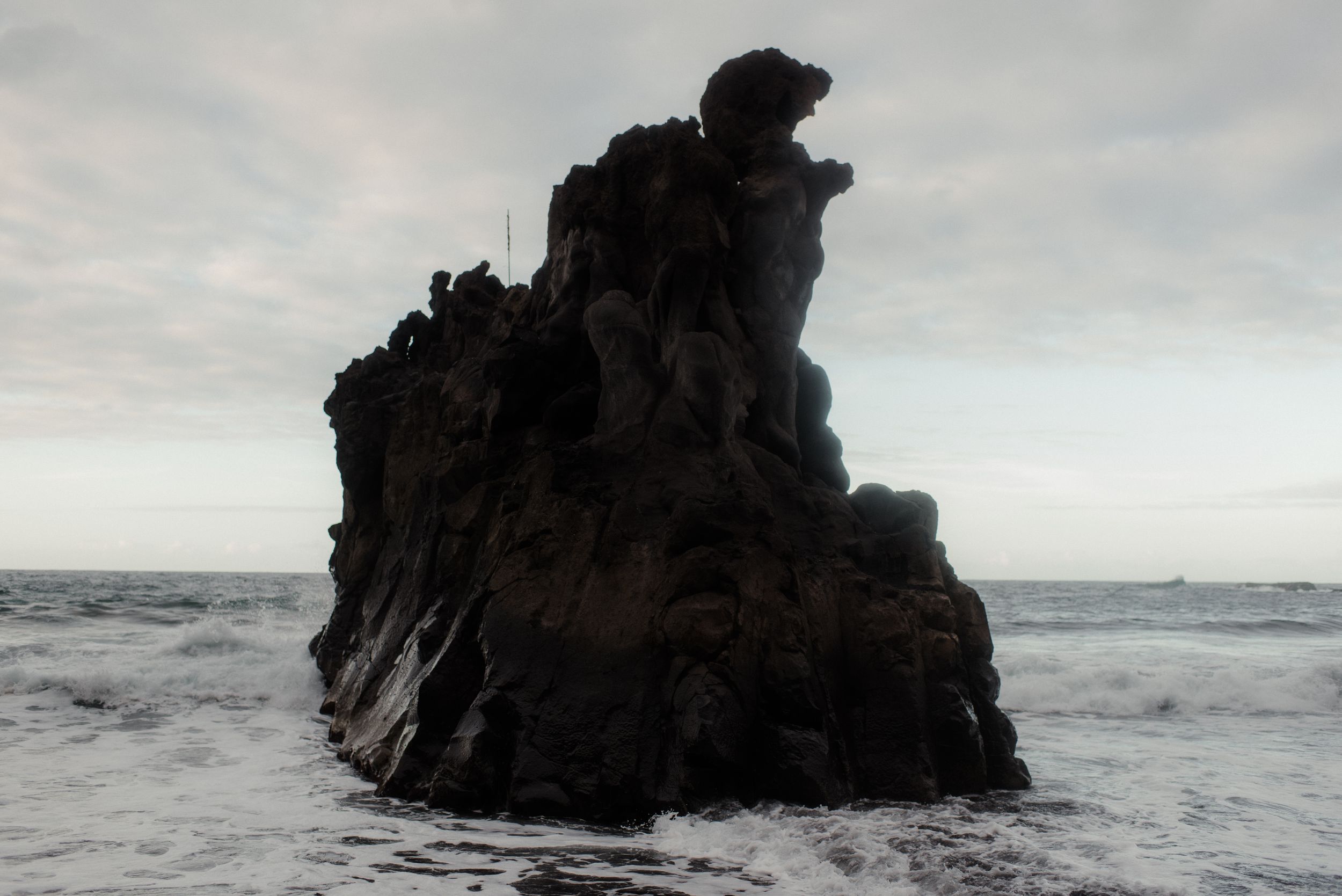 A dramatic black rock formation rises from the churning ocean waters against an overcast sky.