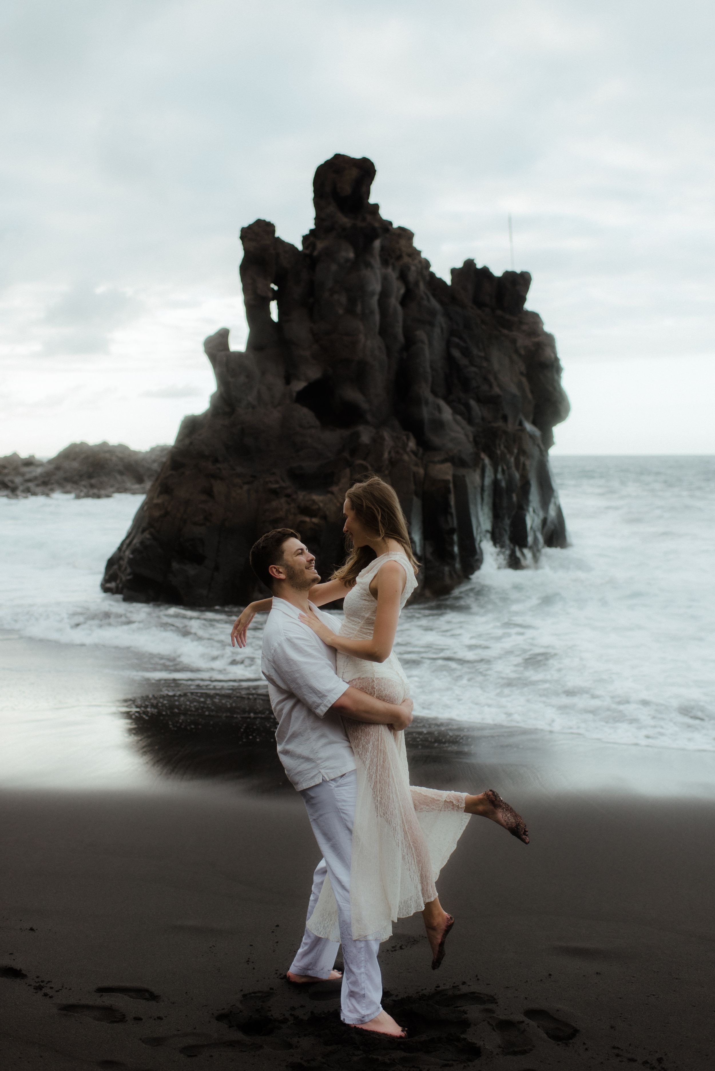 A romantic couple dances together on a black sand beach with dramatic rocky cliffs and crashing waves in the background.