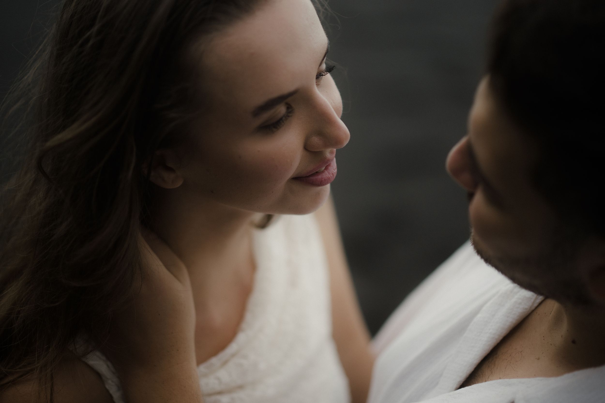 A couple in formal wedding attire share an intimate moment on a rocky beach at sunset.
