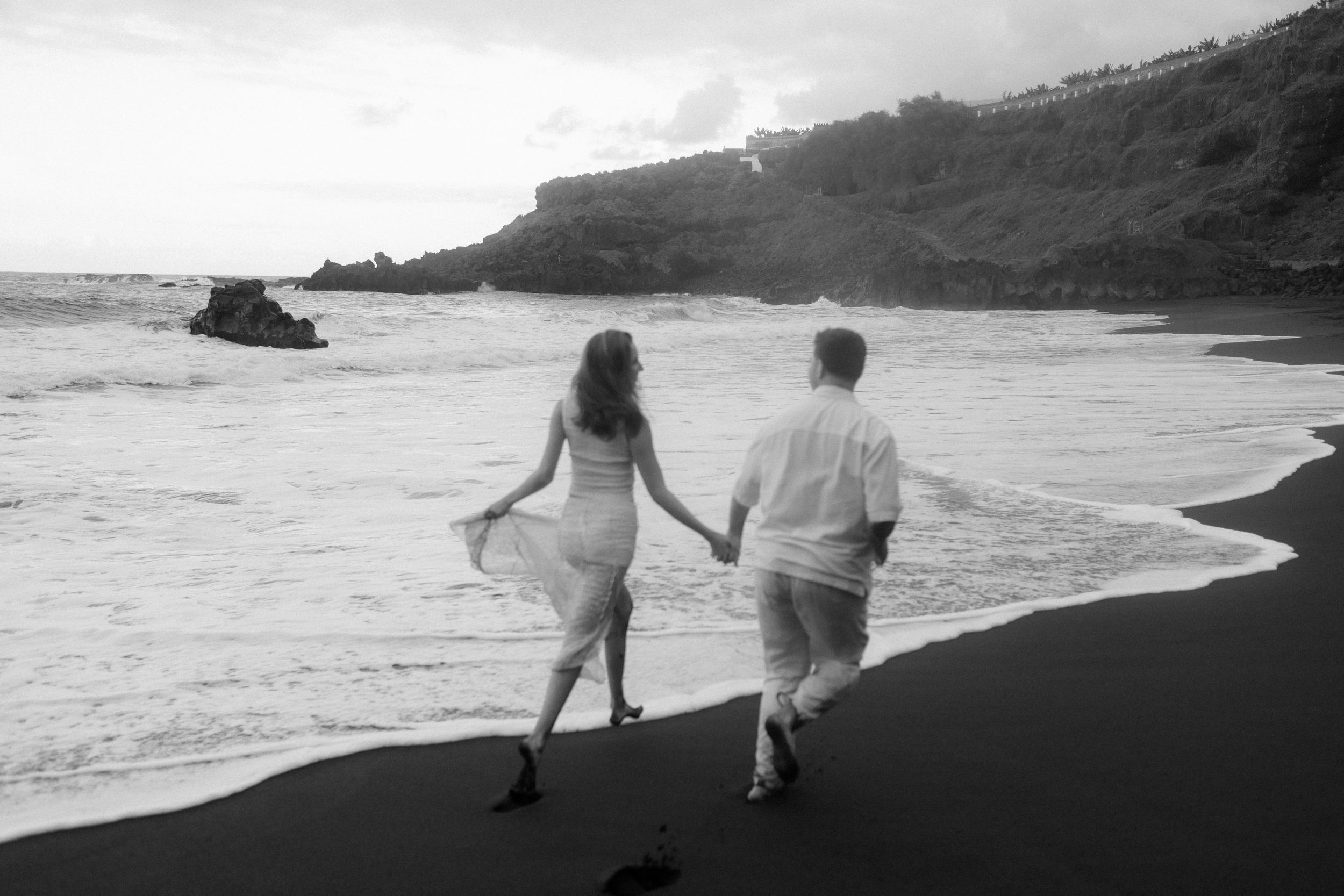 Silhouettes of two people walking along a dramatic rocky coastline at sunset with waves crashing against cliffs.