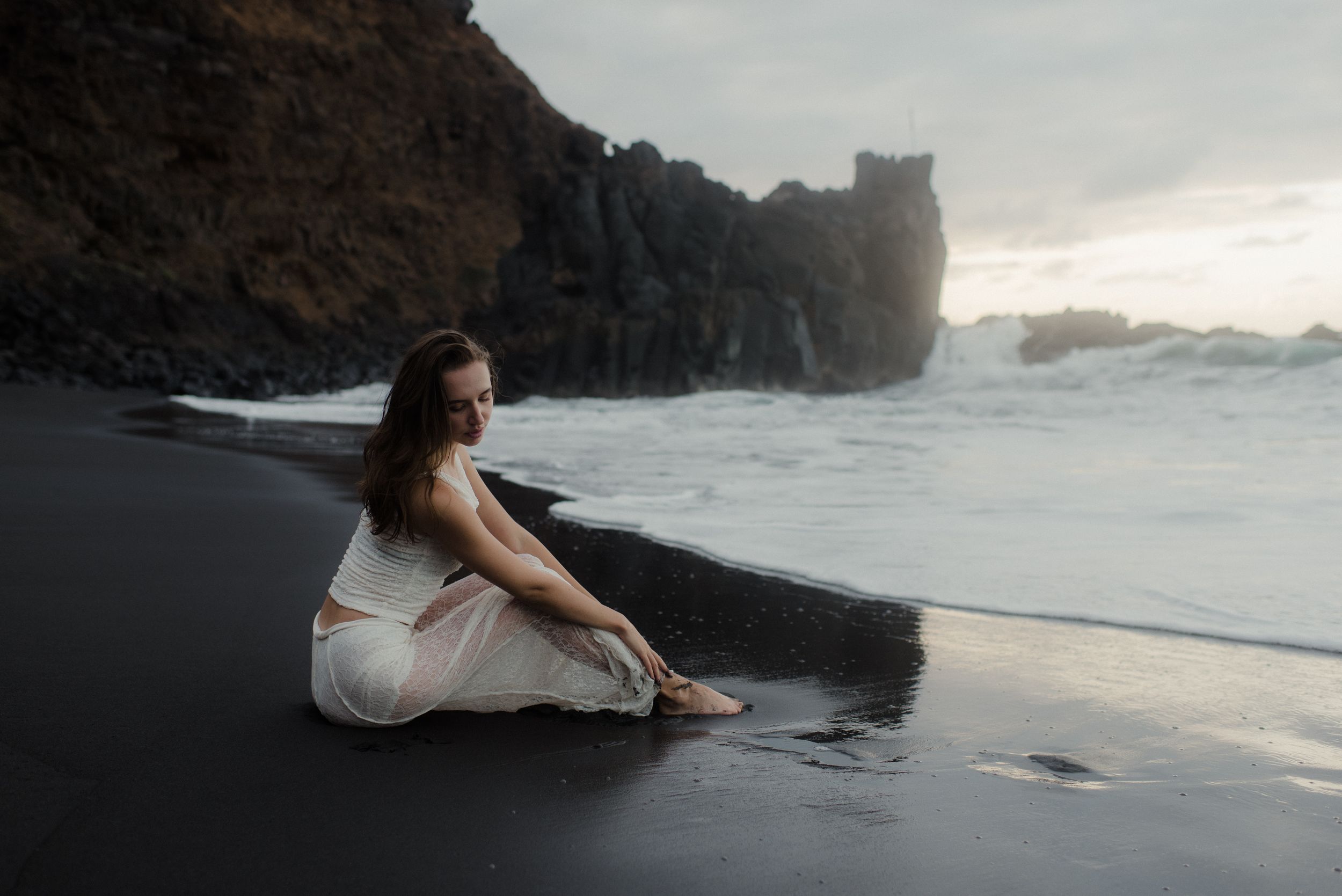 A romantic scene unfolds as a couple in white sits on dark volcanic sand by the ocean.