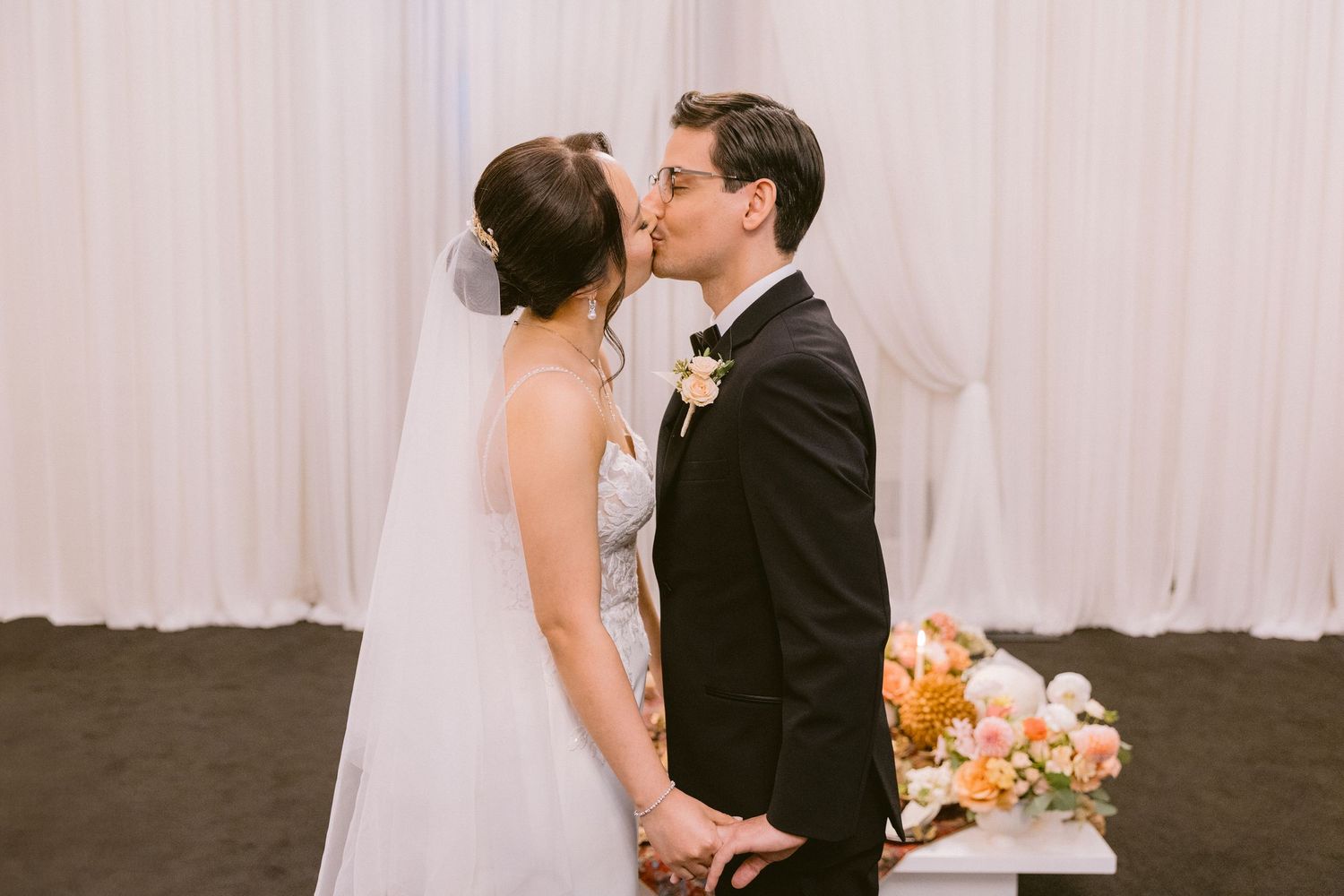 A romantic wedding kiss between newlyweds in formal attire with a bouquet of peach flowers beside them.