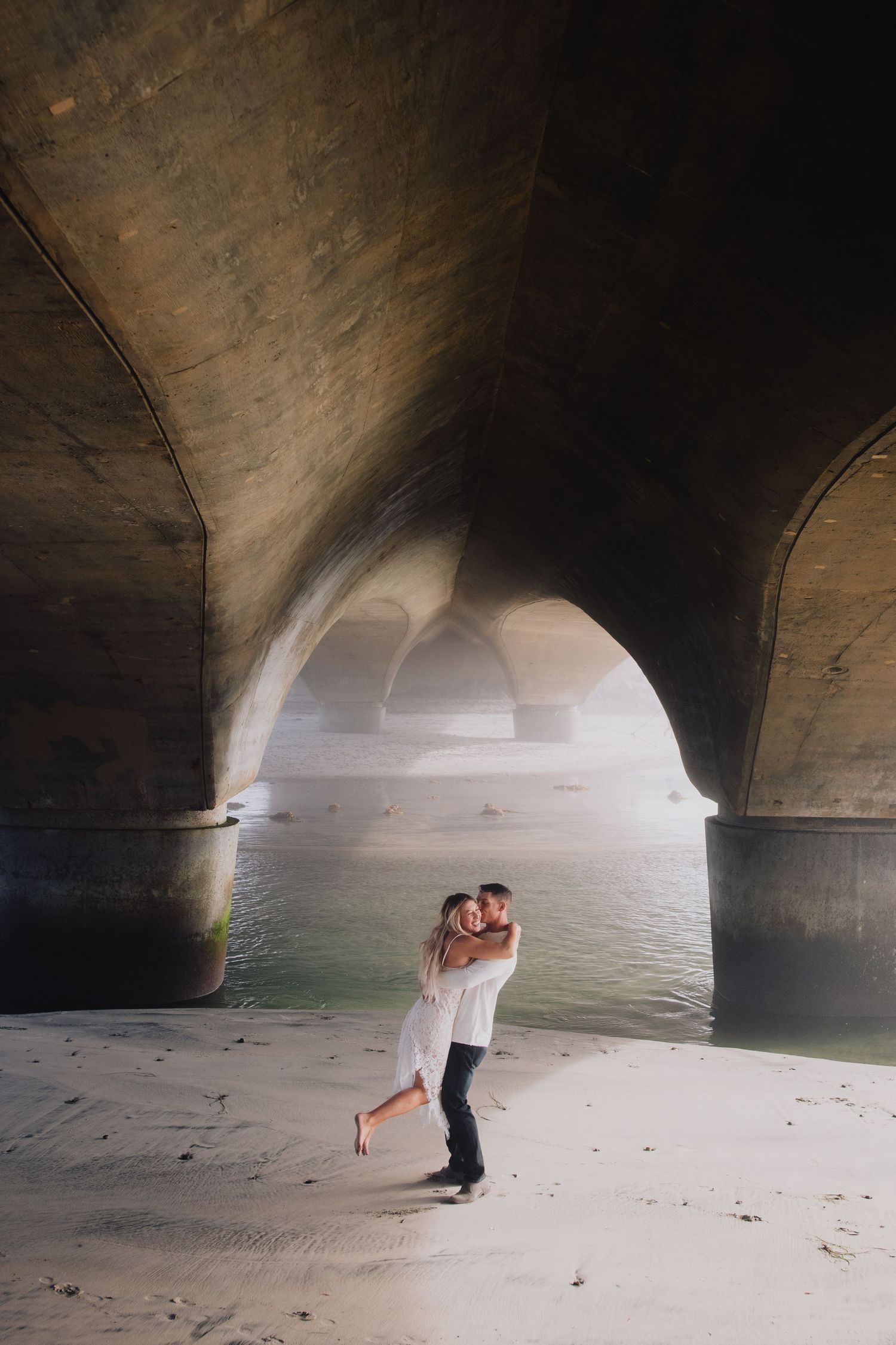 Couple sharing a romantic embrace under a large stone bridge arch near water on a sandy riverbank.