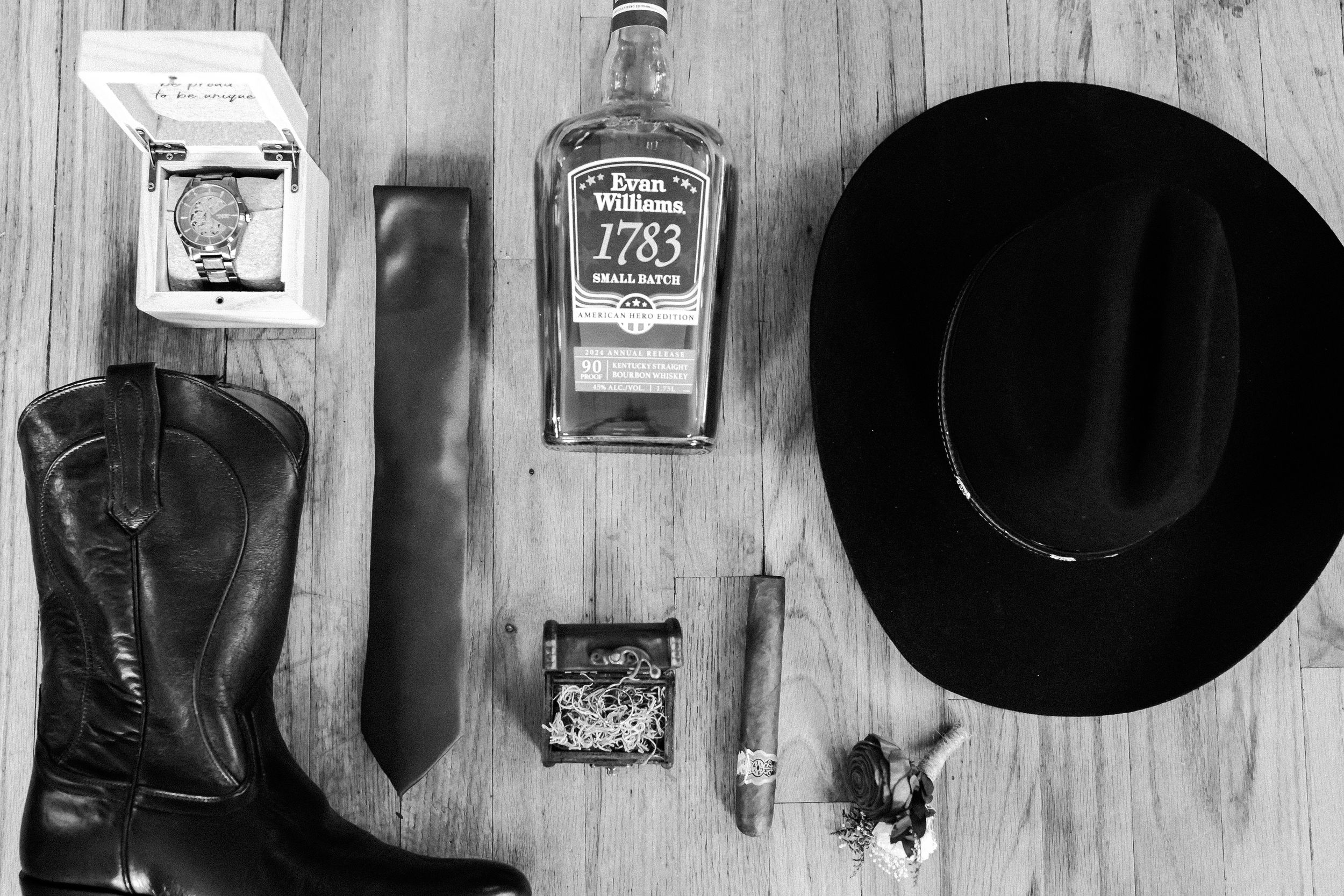 Black and white photo of western boots, knife, Jack Daniels whiskey bottle, black cowboy hat and accessories on wooden floor.