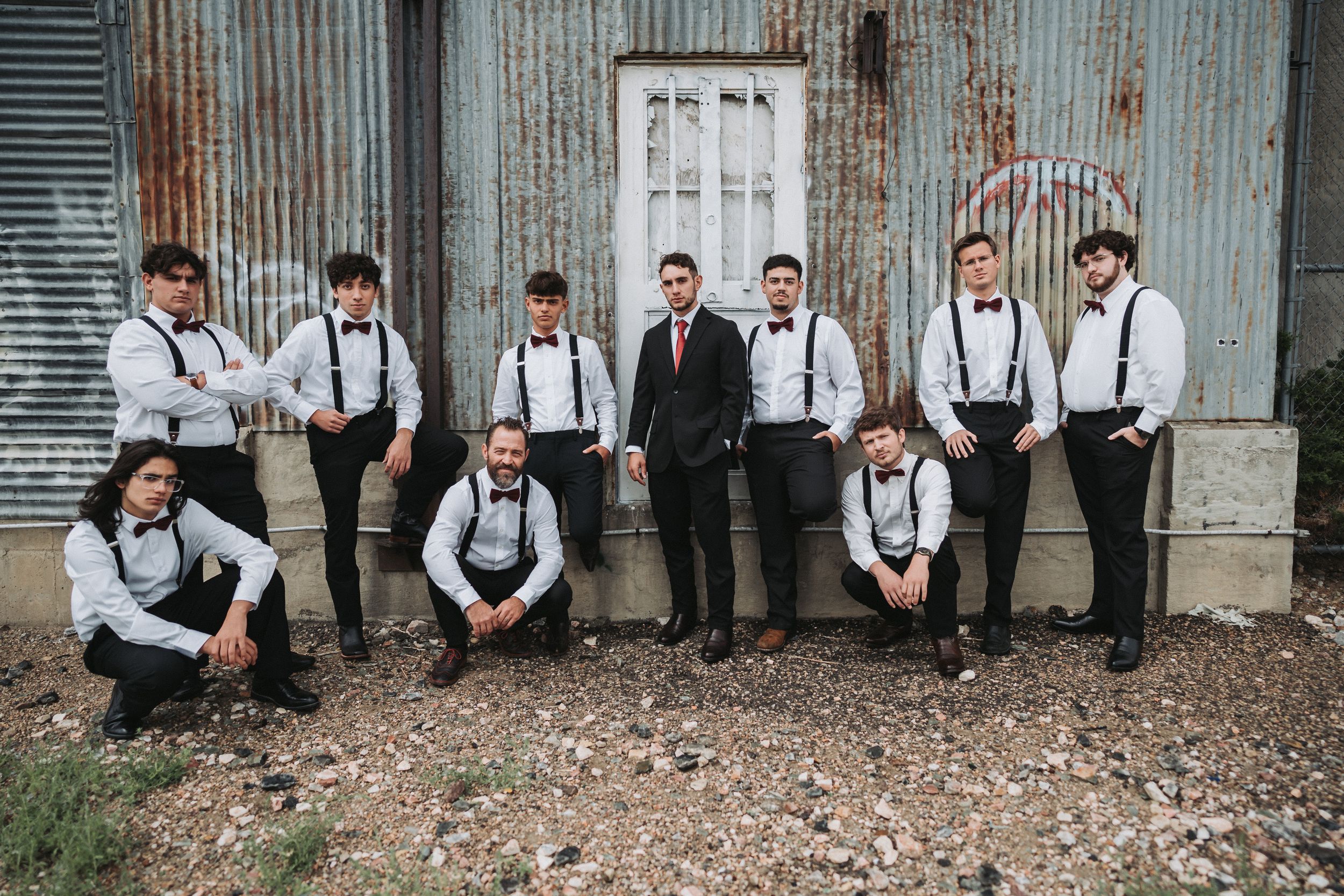 Wedding party in formal attire poses casually against rustic wall in black and white photography series.