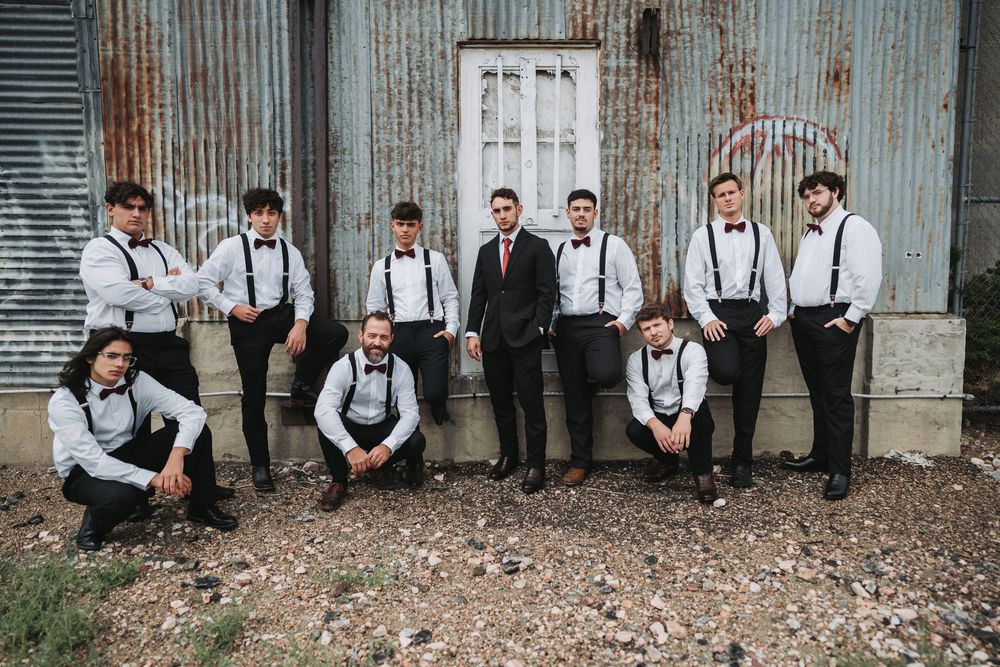 Wedding party in formal attire poses casually against rustic wall in black and white photography series.