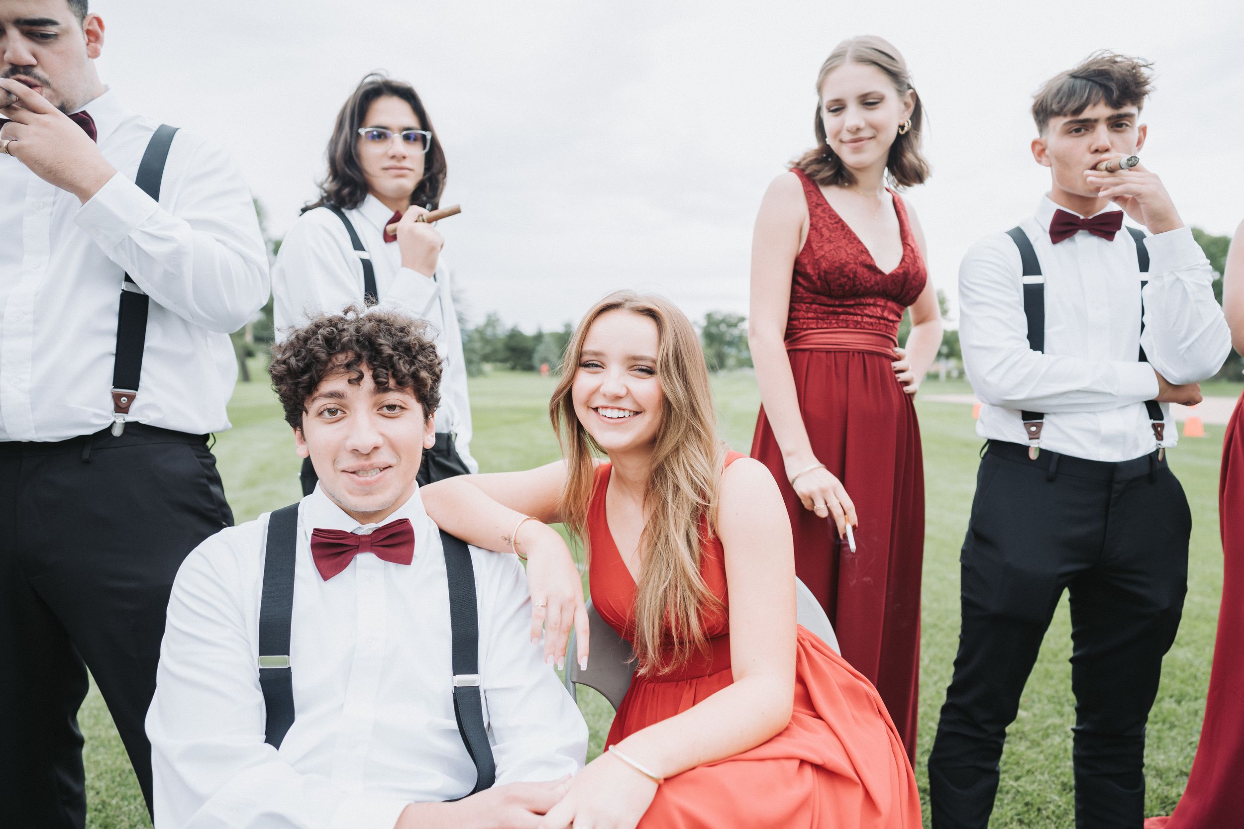 Wedding party members in formal attire pose together outdoors in a mix of red dresses and black suits.