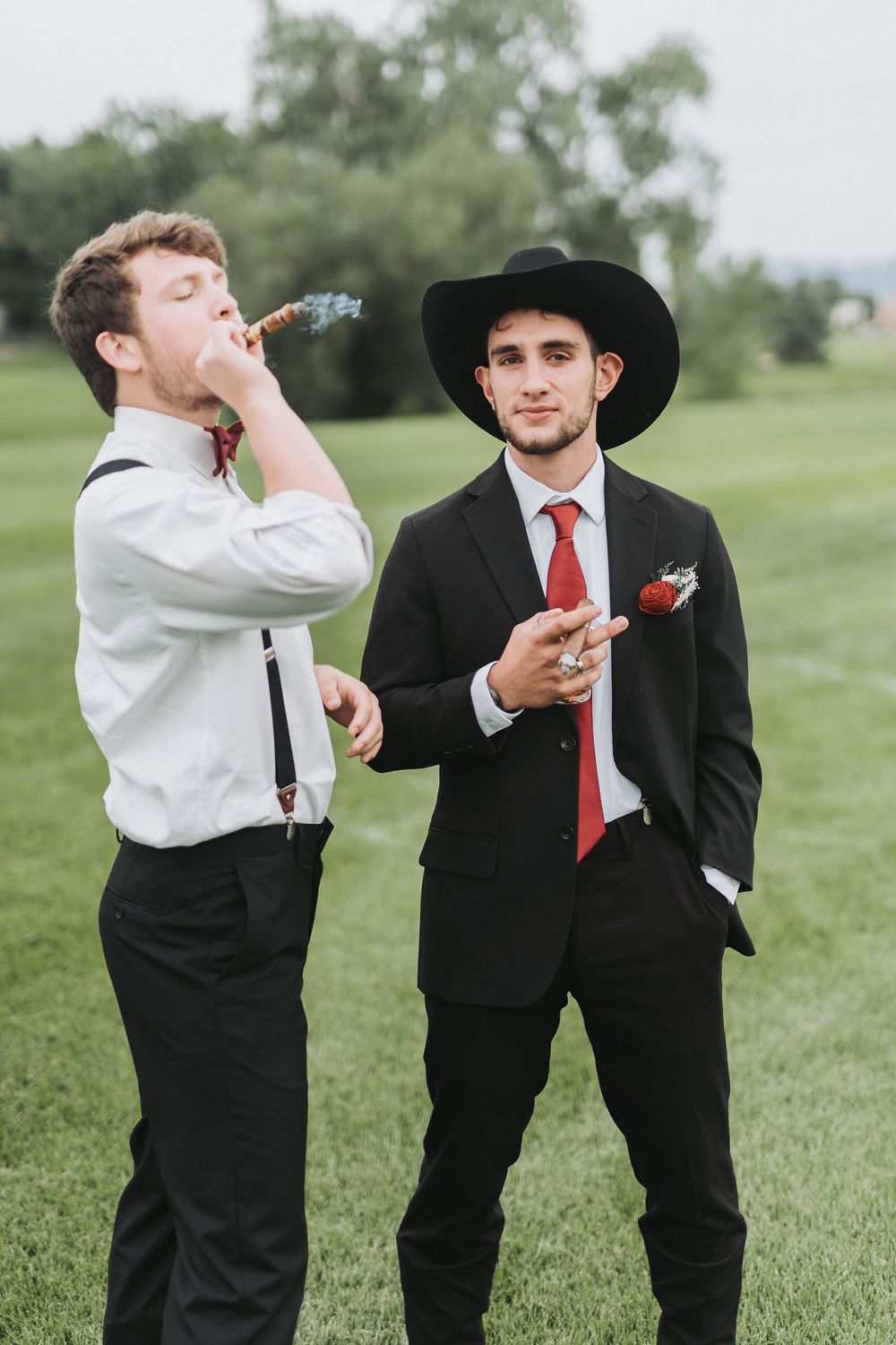 Two people in formal attire playfully taking shots together in a grassy outdoor setting.
