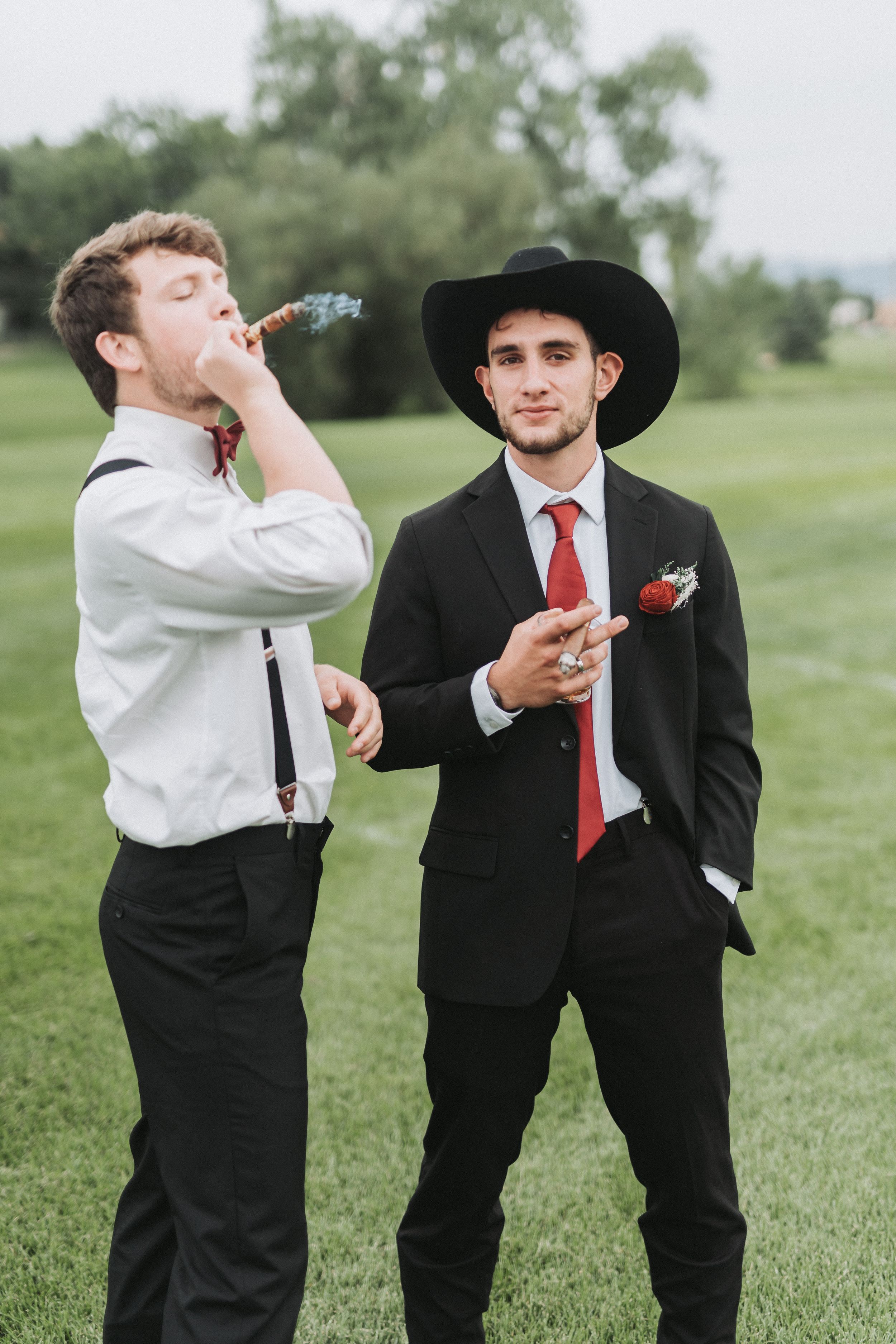 Two people in formal attire playfully taking shots together in a grassy outdoor setting.