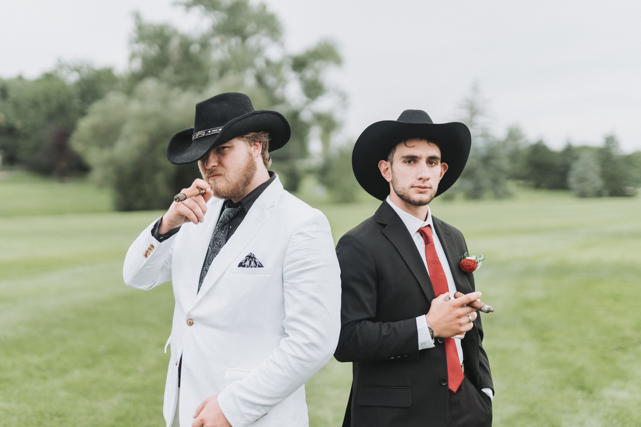 Stylishly dressed wedding party members in black suits and white shirts pose together on a grassy lawn.