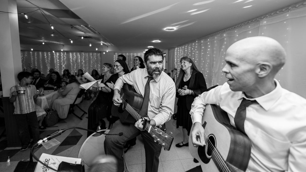Wedding party members dance and celebrate under string lights at an indoor reception venue.