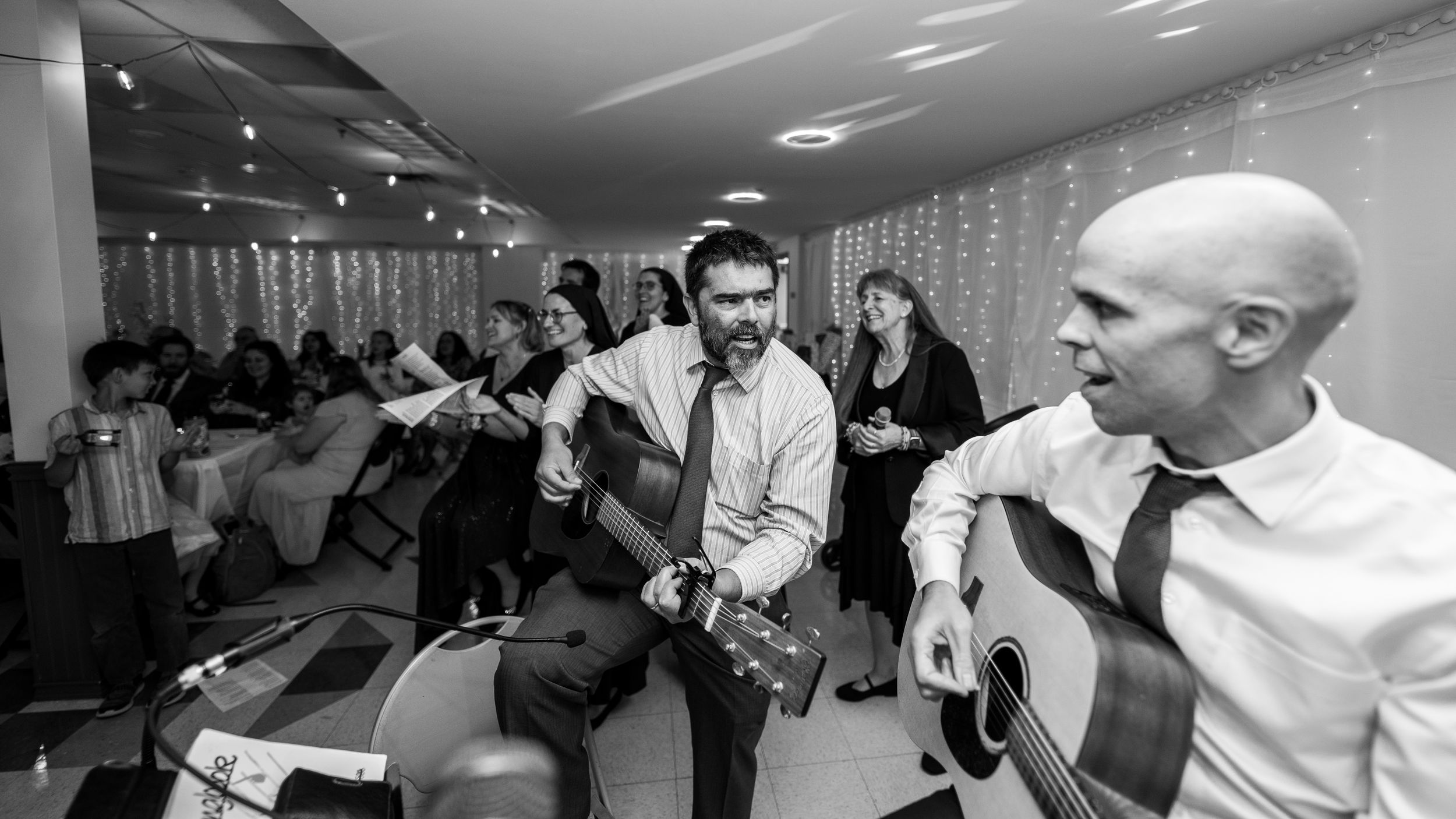 Wedding party members dance and celebrate under string lights at an indoor reception venue.