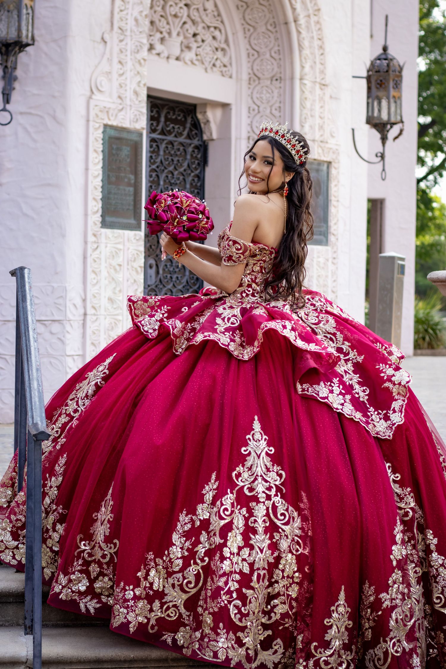 Bride in ornate red and white lace ballgown holds bouquet in front of decorative archway.