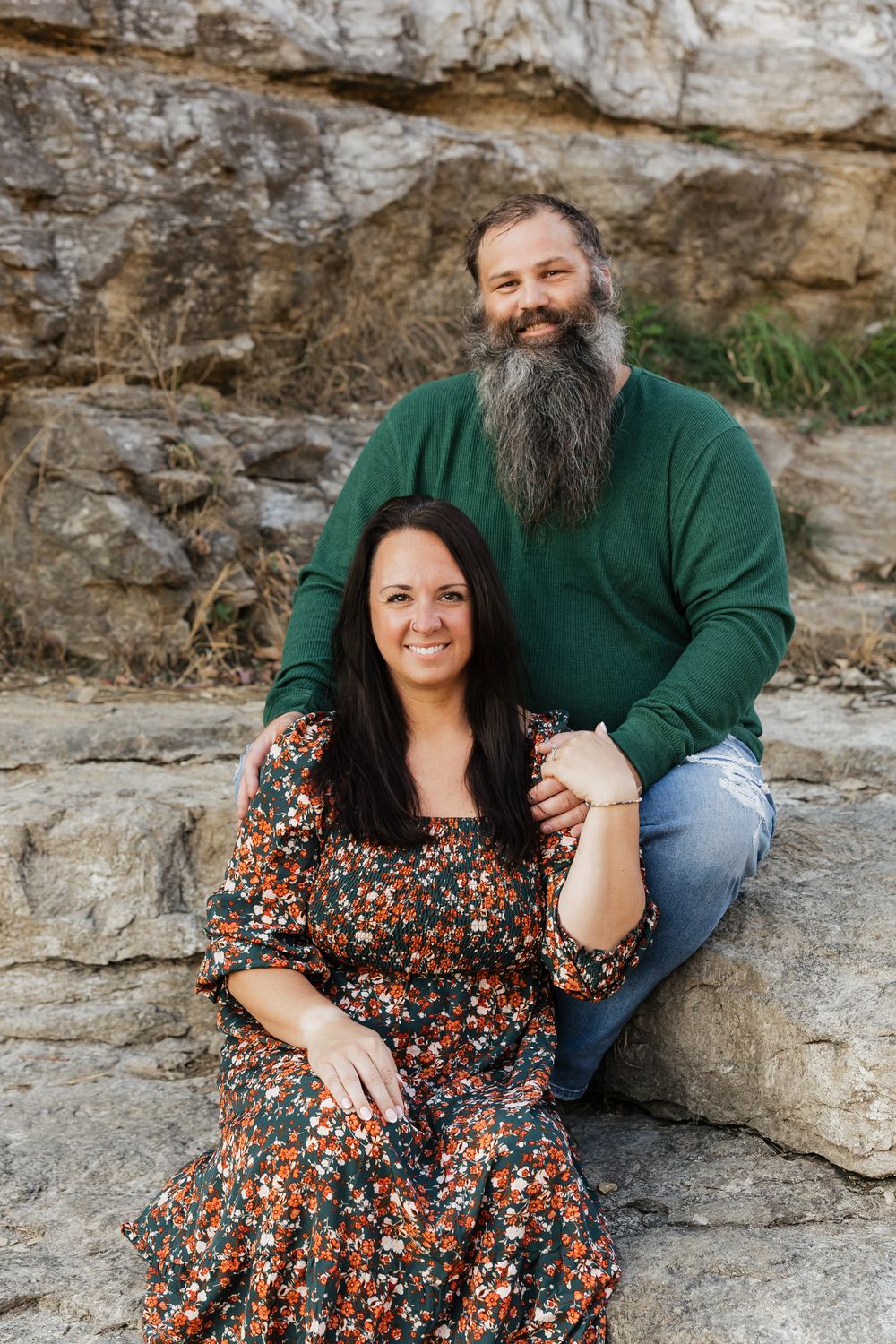 A couple sitting together on rocky terrain, with the person in green wearing a long beard.
