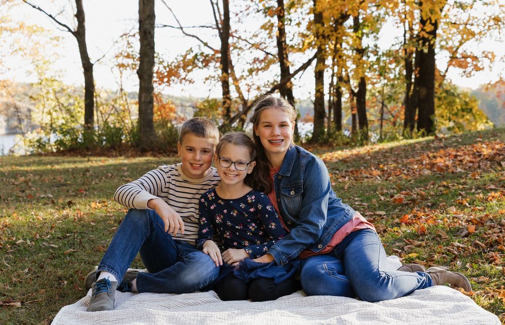 A family relaxes together on a white blanket surrounded by colorful autumn leaves in a park setting.